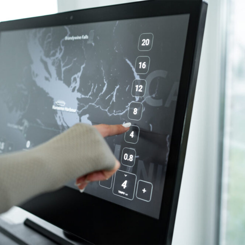 A finger touches a treadmill screen displaying a map and numbers. The screen is on a black treadmill.