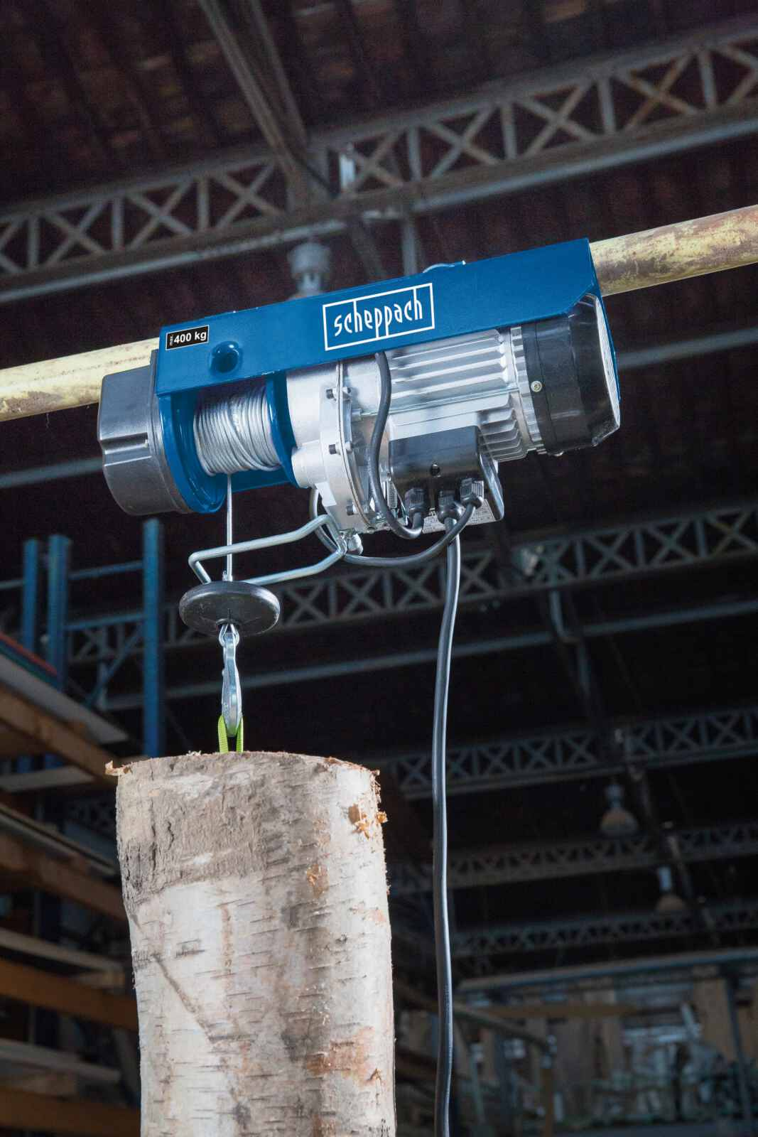Paranco elettrico blu e argento che solleva un tronco. Il paranco è sospeso a una struttura metallica.