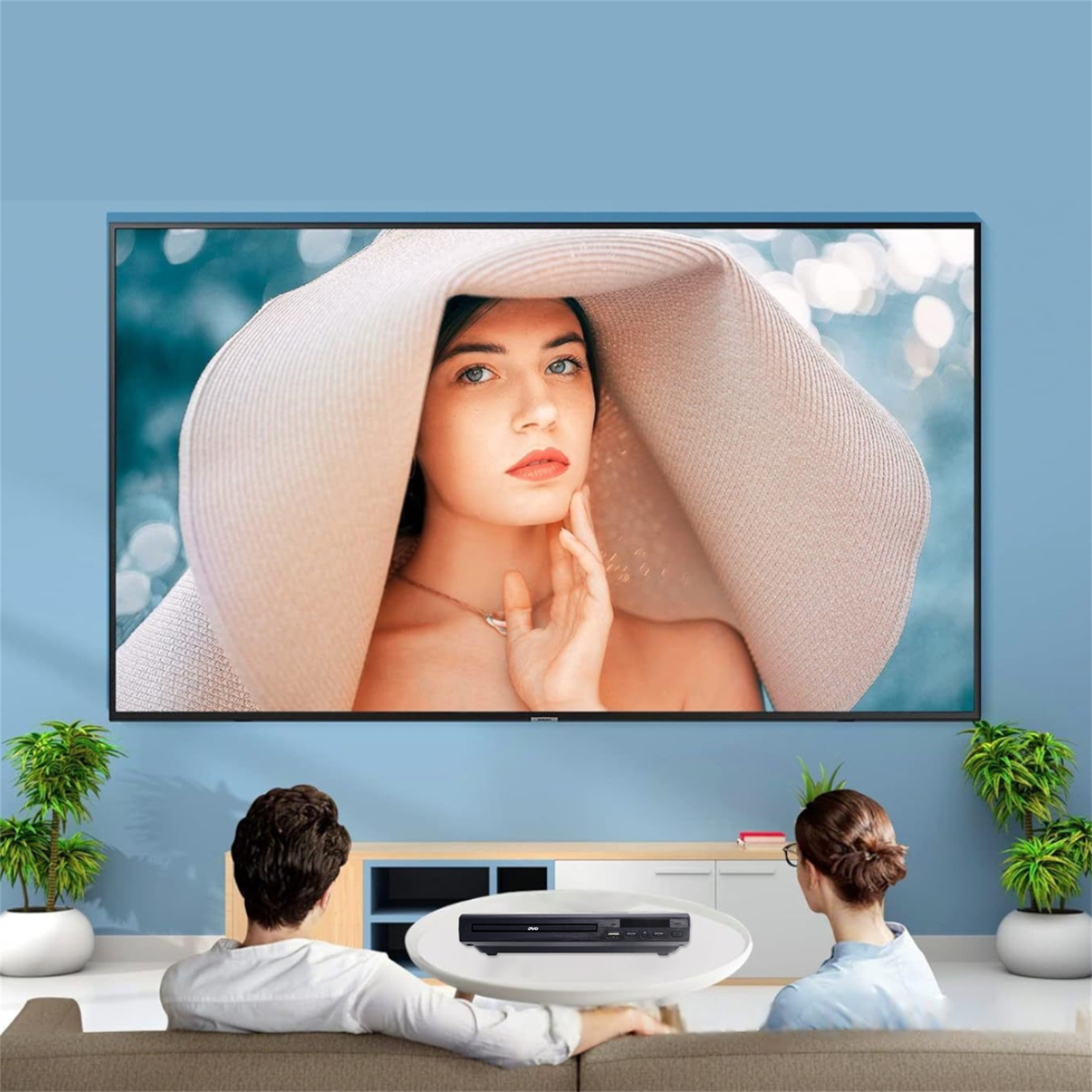 Zwei Personen sehen fern. TV zeigt Frau mit Hut. DVD-Player steht auf dem Tisch.