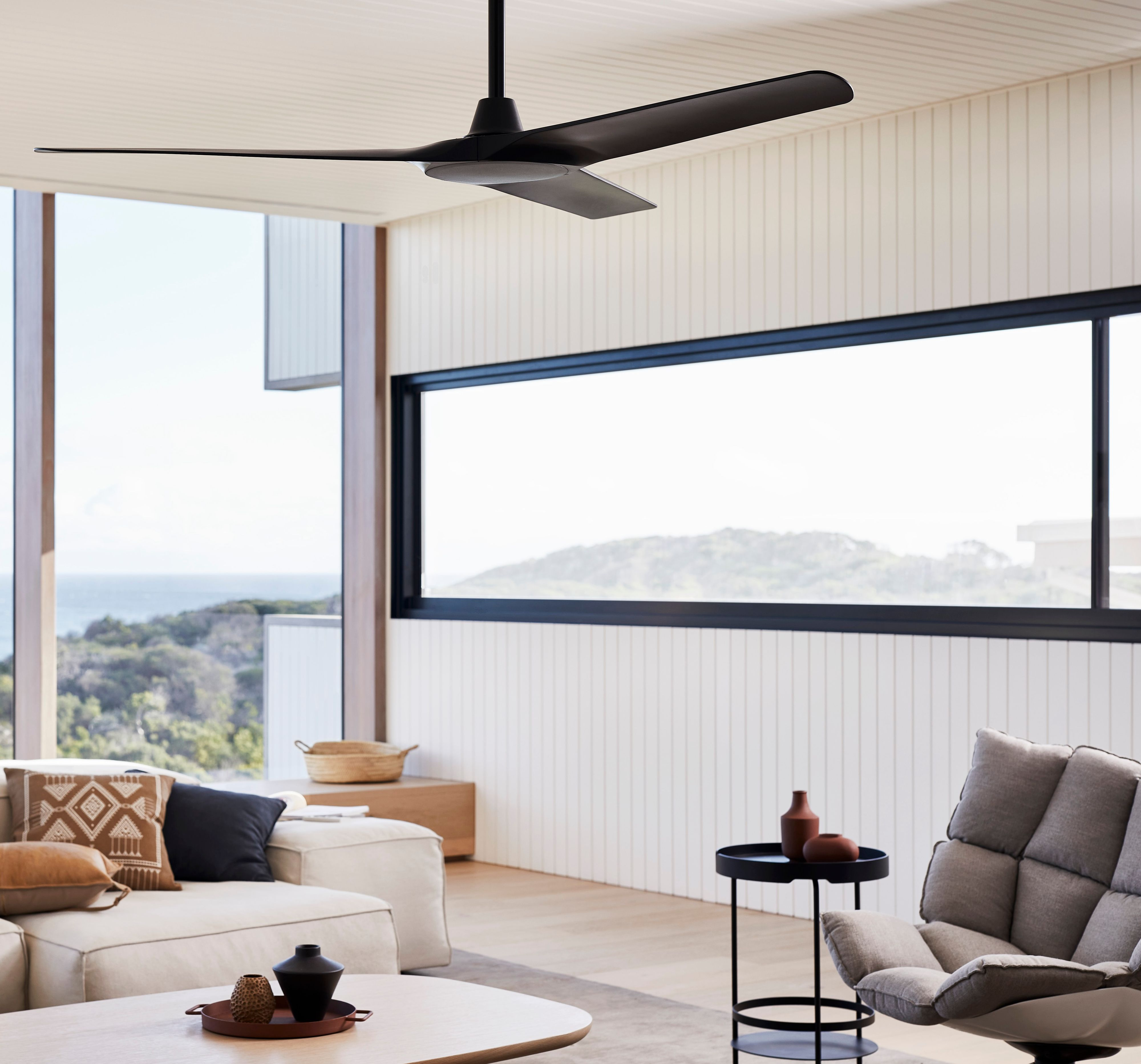 Modernes Wohnzimmer mit großen Fenstern, Deckenventilator, Sofa, Sessel und Blick auf Meer und Landschaft.