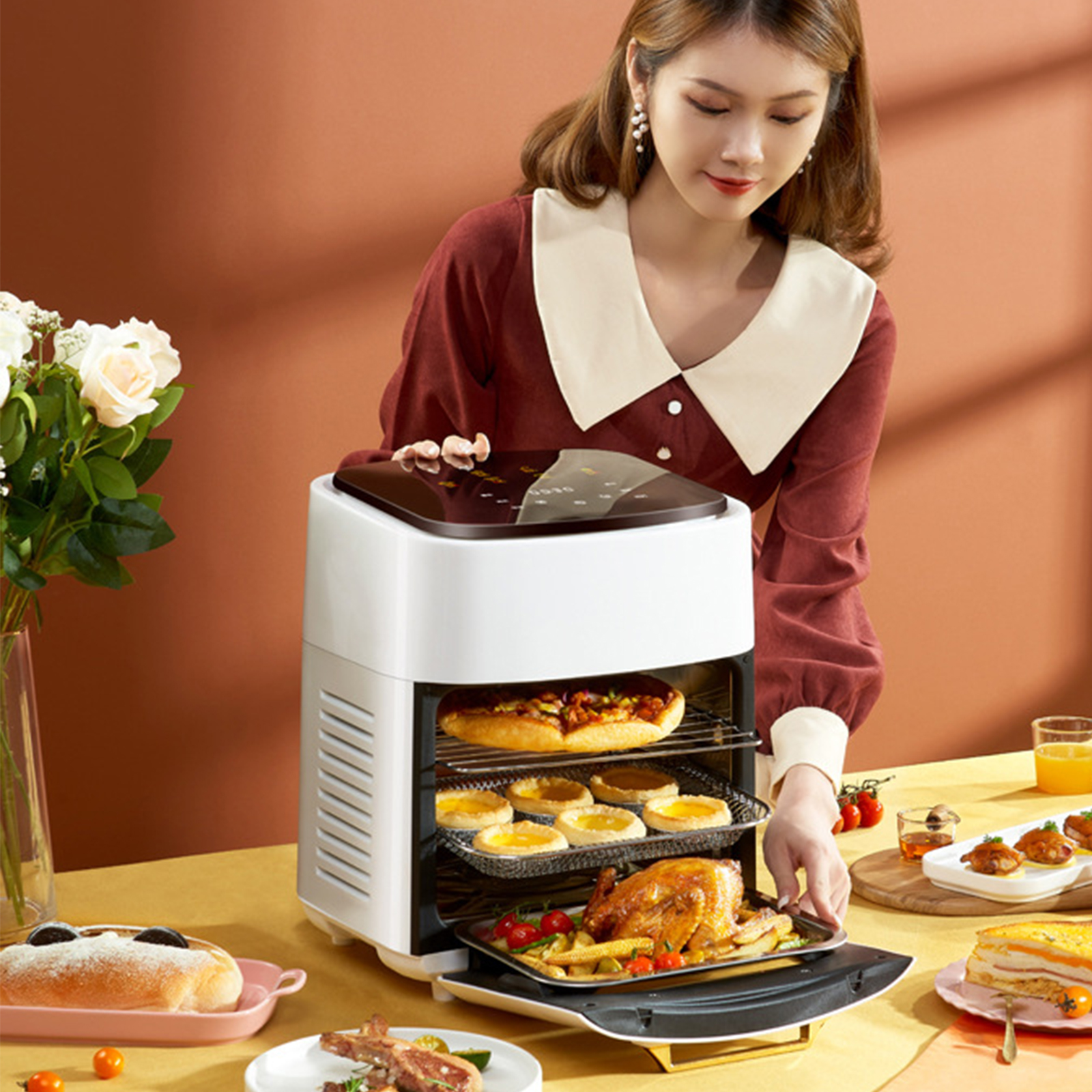 Eine Frau mit Essen neben einer Heißluftfritteuse. Pizza, Gebäck und ein Brathähnchen sind sichtbar.