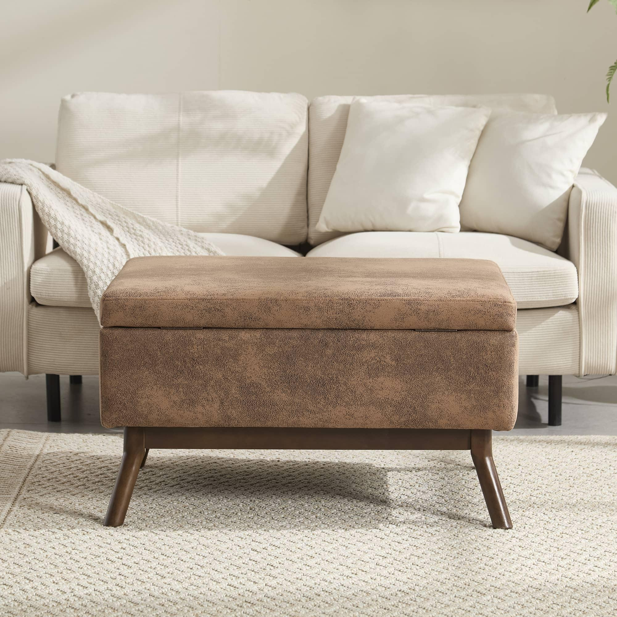 Eine braune Aufbewahrungshocker steht vor einem weißen Sofa. Der Hocker hat einen Deckel und steht auf Holzbeinen.
