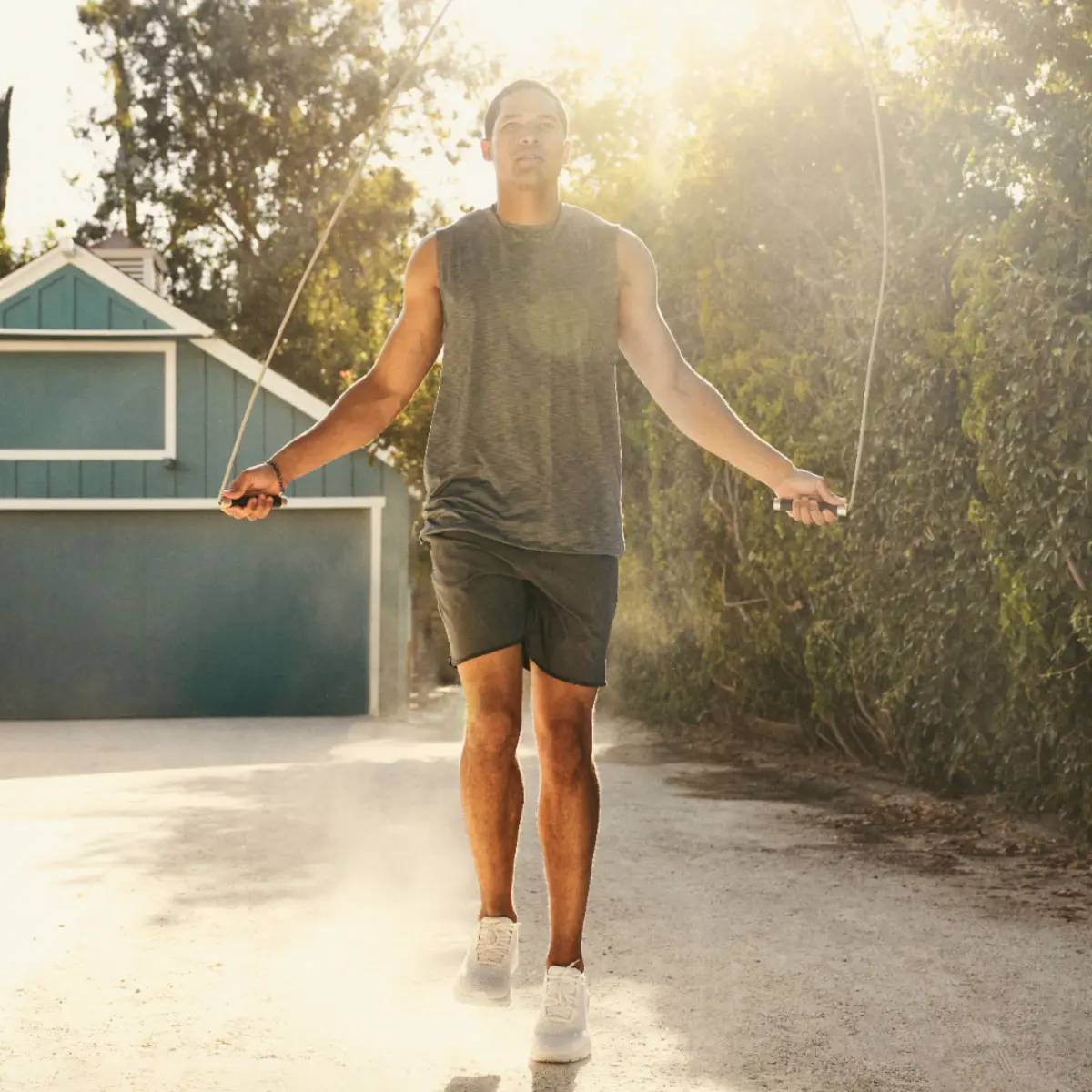 Mann springt Seil auf einem Feldweg. Graues Tanktop, schwarze Shorts, weiße Schuhe. Sonniger Hintergrund.