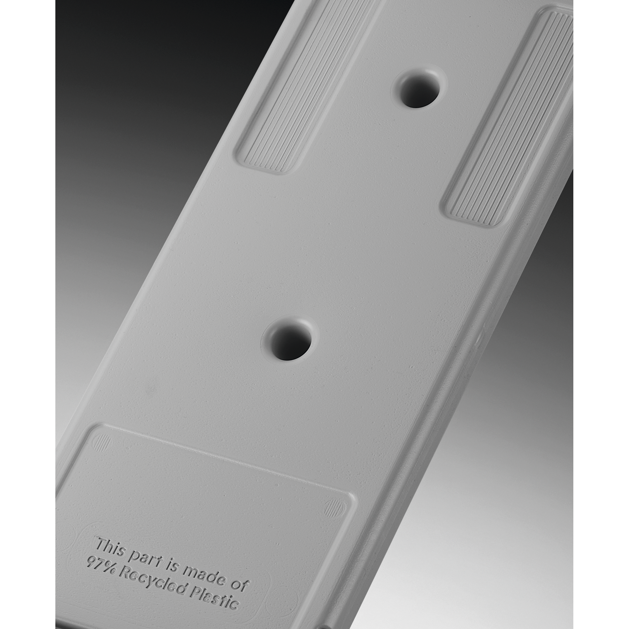 Primer plano de una pieza de plástico gris con dos agujeros y texto: 'This part is made of 97% Recycled Plastic'.