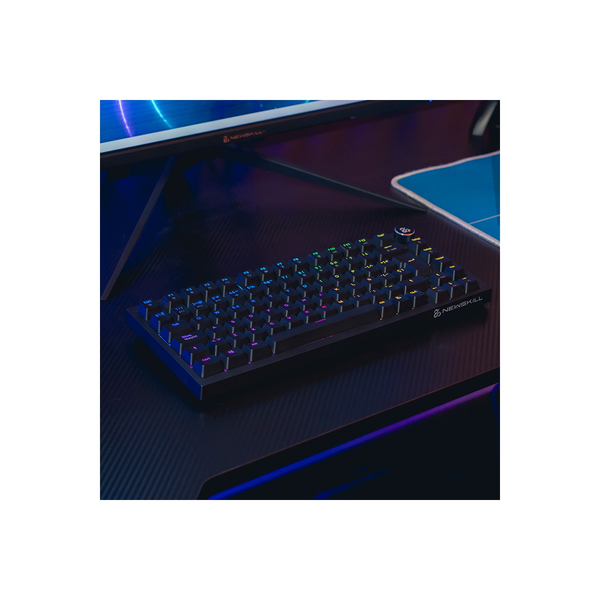 Tastiera meccanica nera illuminata con più colori, su un tavolo scuro. Un monitor sullo sfondo.