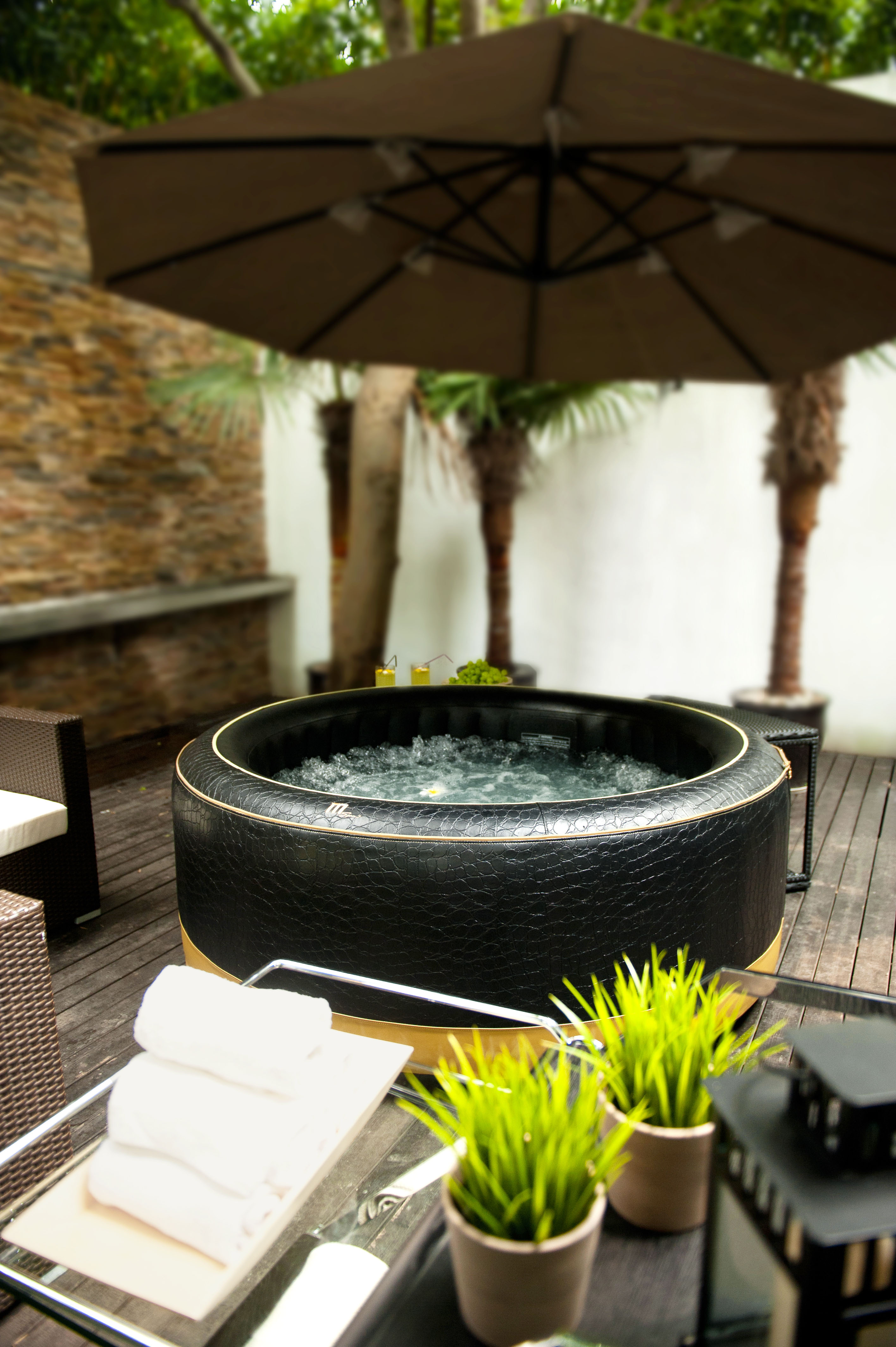 Outdoor-Szene mit schwarzem Whirlpool, Handtüchern und Pflanzen. Palmen und ein großer Sonnenschirm im Hintergrund.