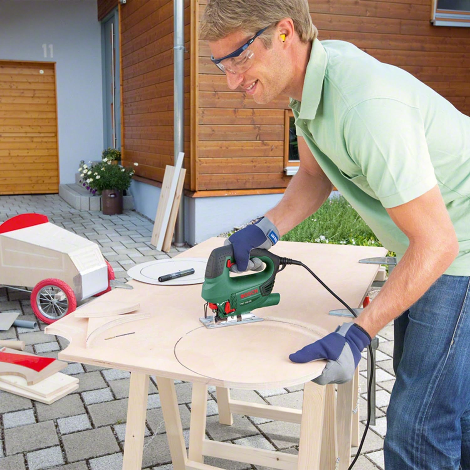 Mann benutzt eine Stichsäge auf Holz. Ein Wagen ist im Hintergrund. Trägt Schutzbrille und Handschuhe.