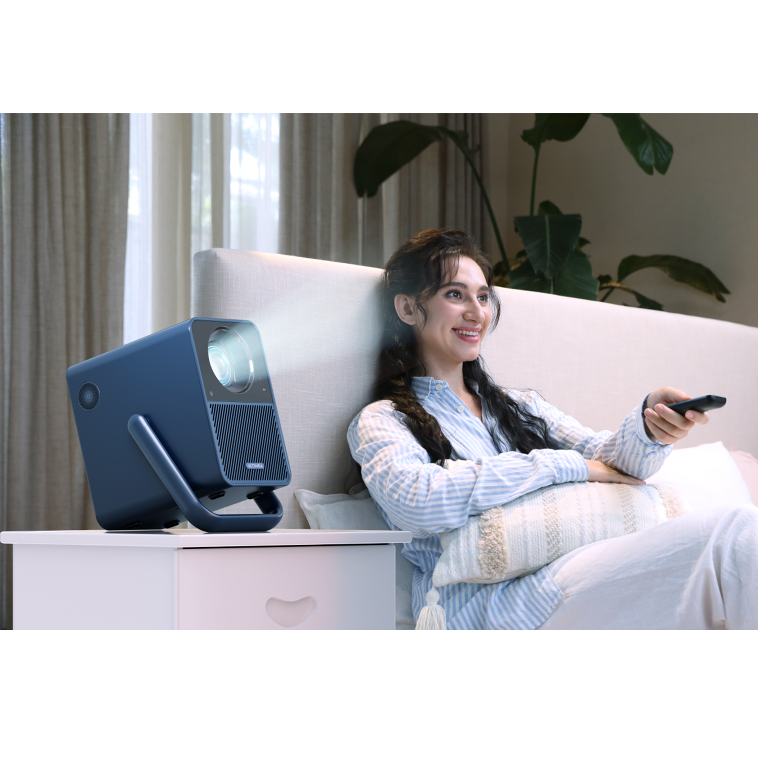A woman watches a movie on a projector. The projector is blue, and on a white nightstand.