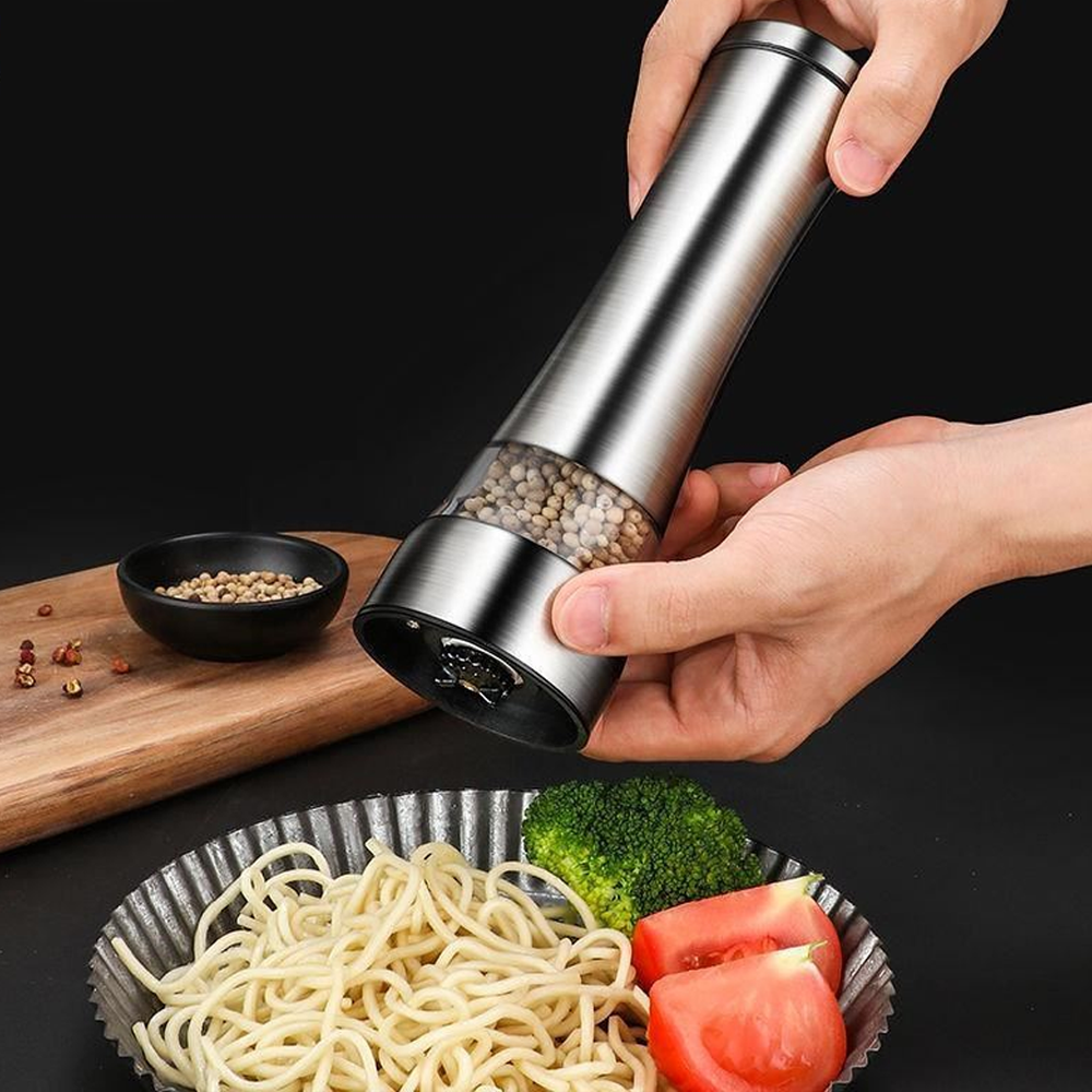 Hände einer Person halten eine silberne elektrische Pfeffermühle über Essen, darunter Nudeln, Brokkoli und Tomaten.