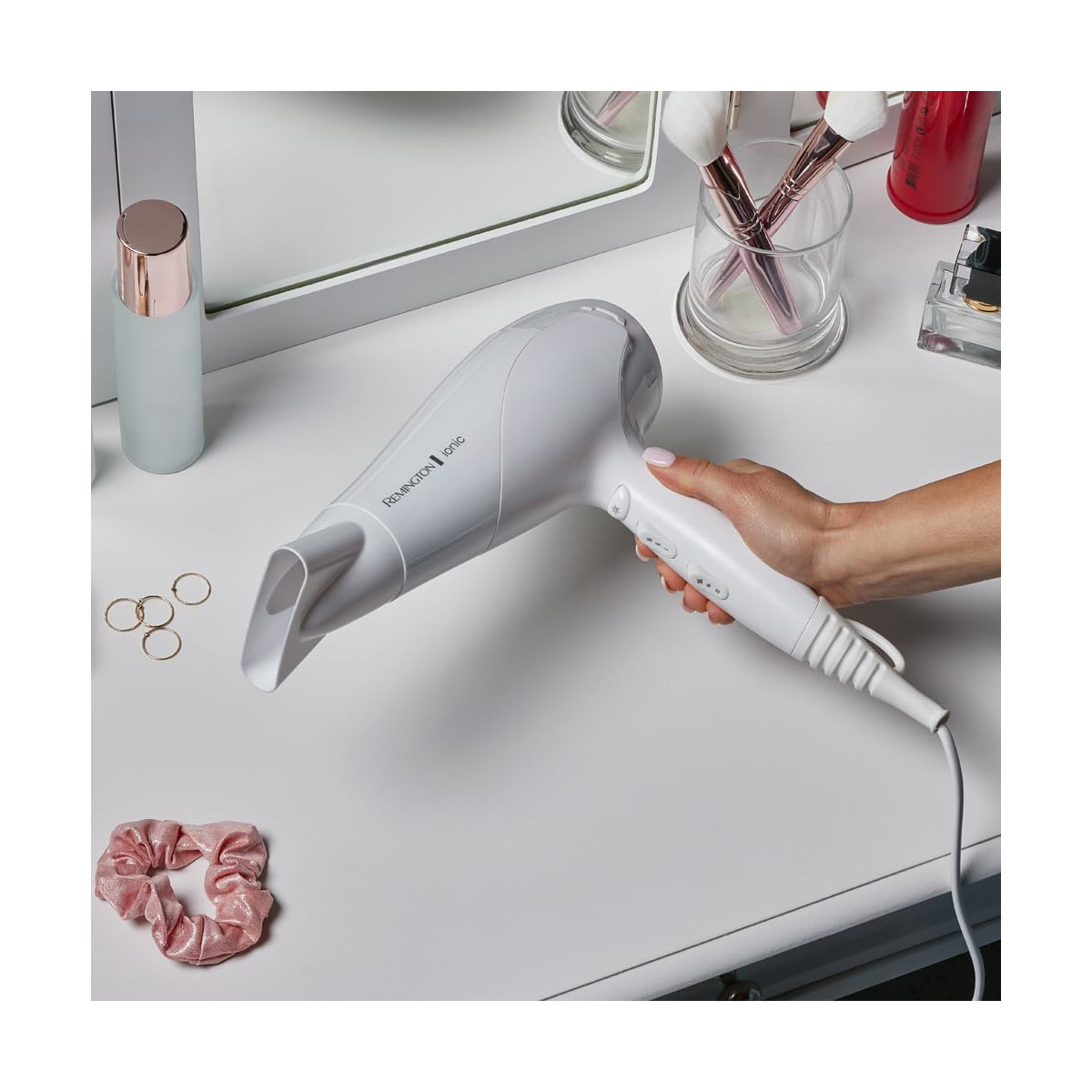 Eine Hand, die einen weißen Haartrockner auf einem weißen Tisch hält. Weitere Gegenstände sind Accessoires.