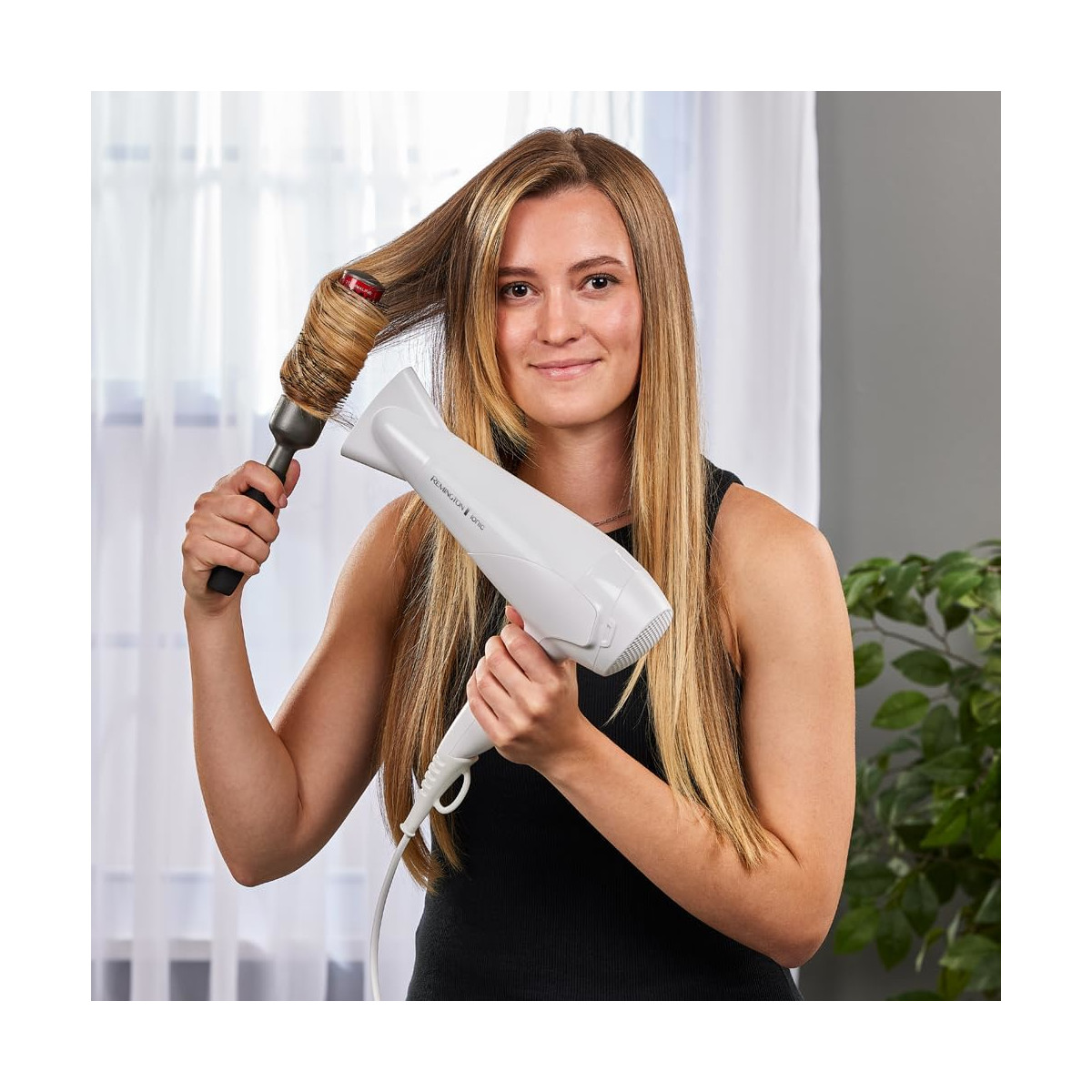 Eine Frau mit blonden Haaren benutzt einen Haartrockner und eine Bürste. Sie schaut in die Kamera und lächelt.