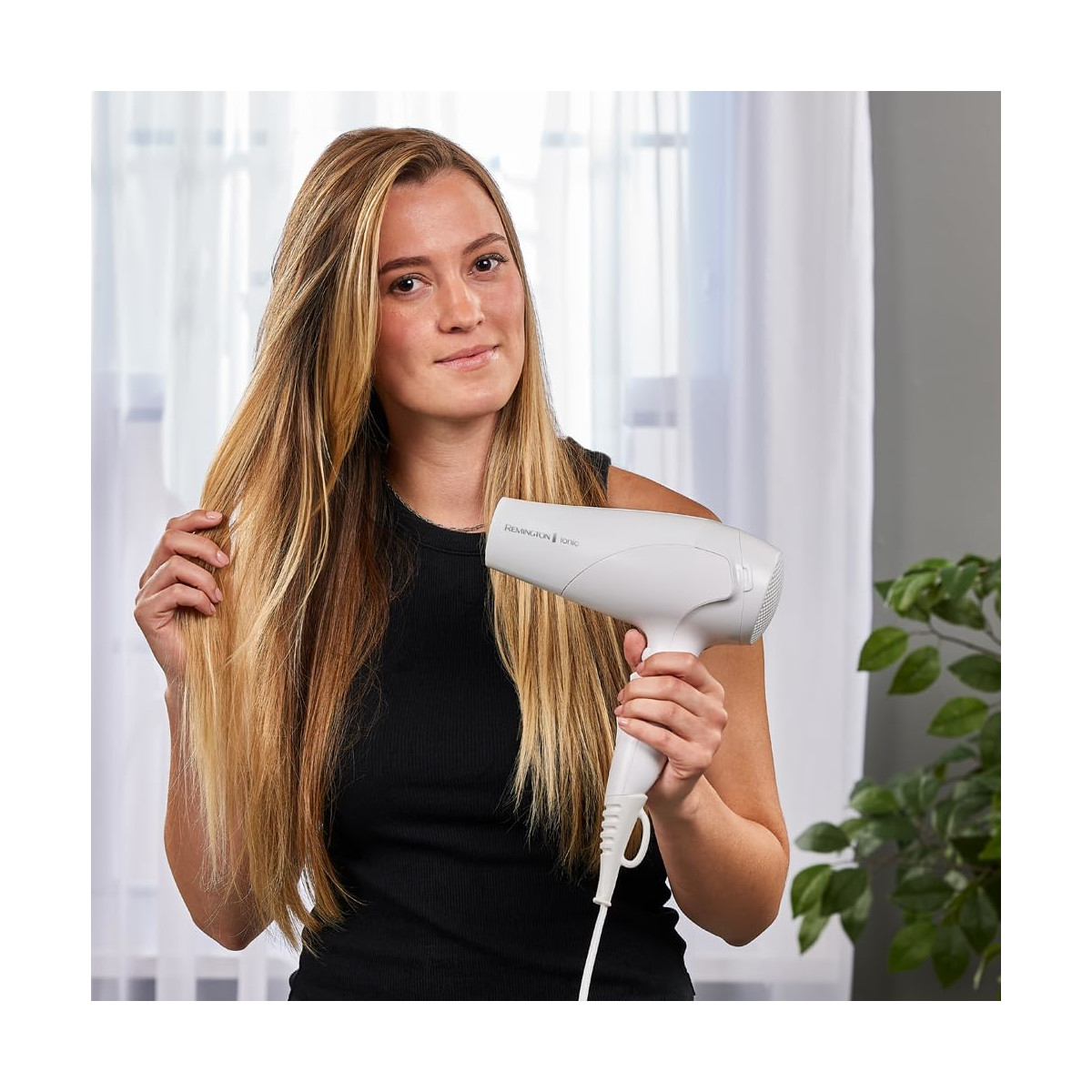 Eine blonde Frau hält einen weißen Haartrockner, ihr Haar ist frisiert und eine Pflanze im Hintergrund.