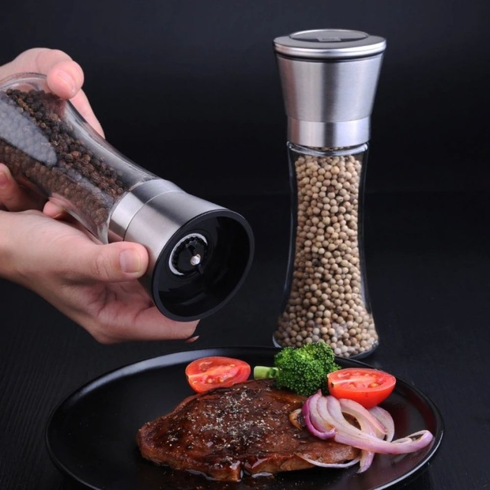 Eine Person verwendet eine Mühle, um Lebensmittel auf einem schwarzen Teller zu würzen. Ein Steak, Gemüse und weiße Pfefferkörner.