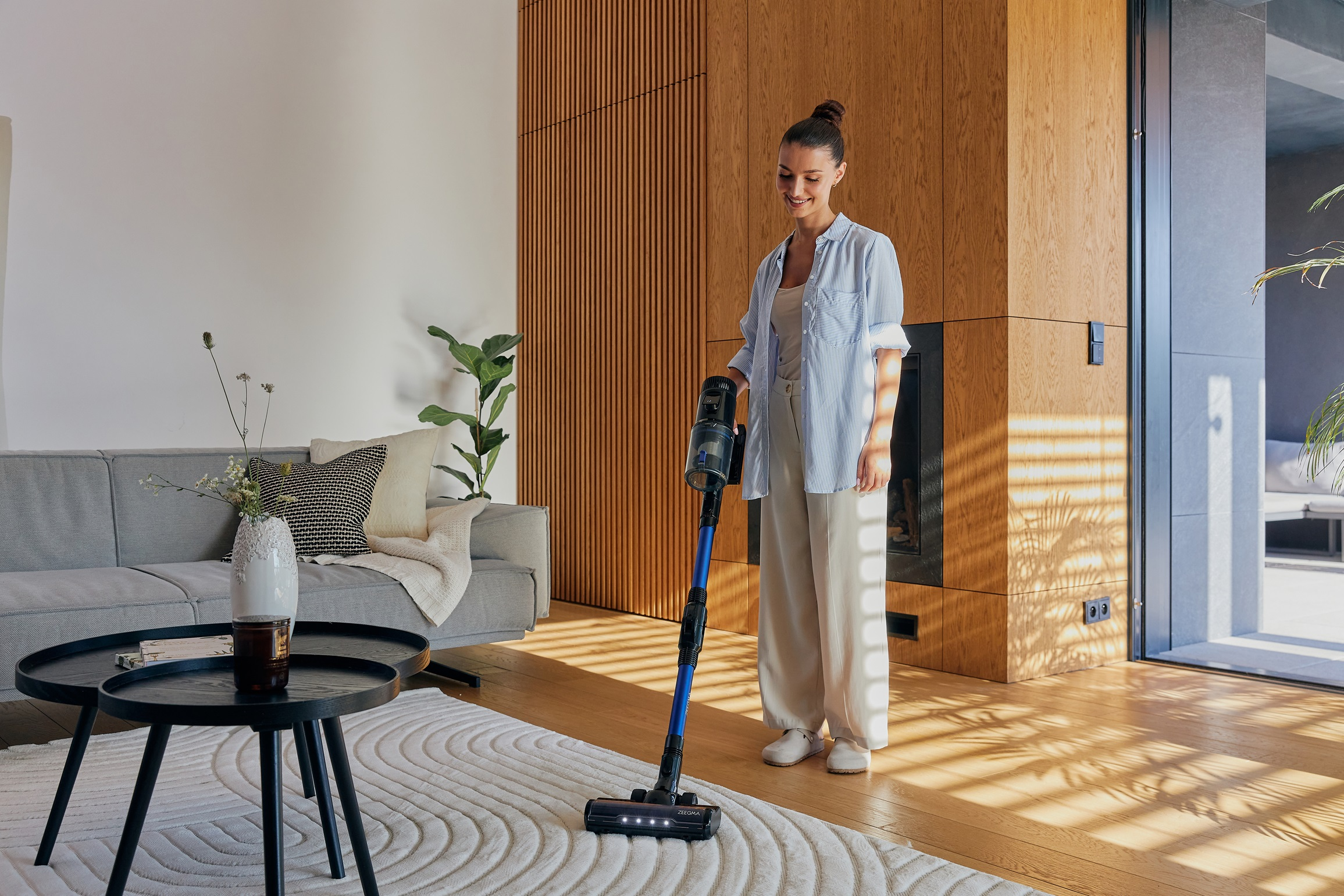 Kobieta odkurza salon. W pokoju znajduje się sofa, stoliki kawowe i drewniane panele na ścianie.