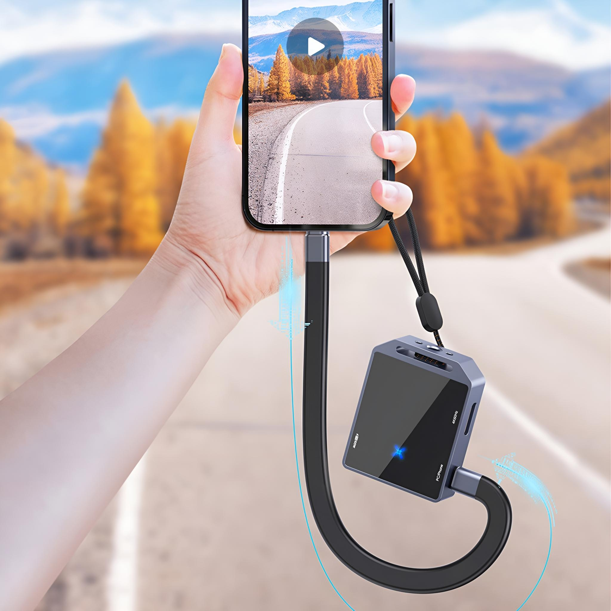 Eine Person hält ein Telefon, das mit einem schwarzen Gerät mit Kabeln verbunden ist. Der Hintergrund zeigt eine Straße und Bäume.