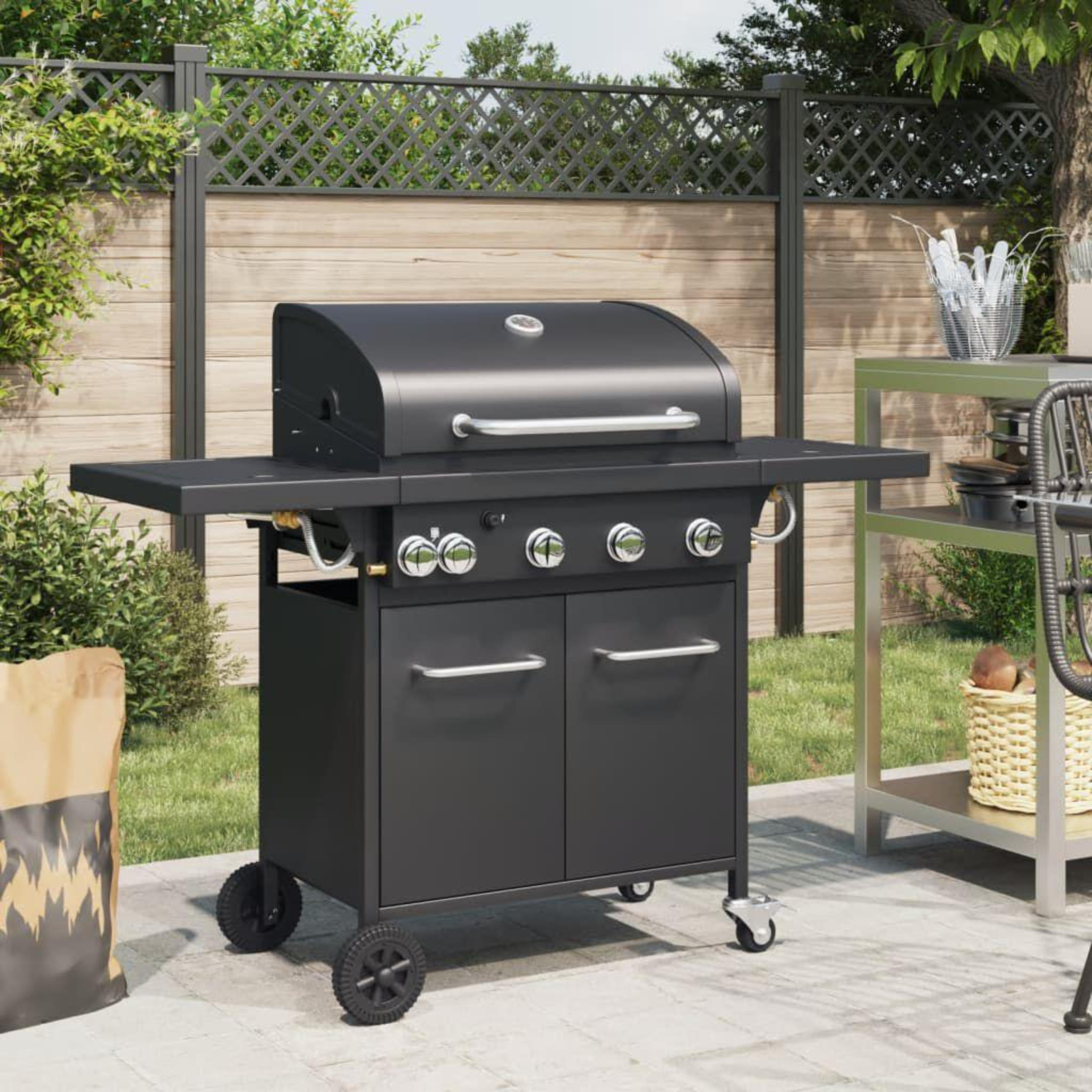 Schwarzer Außengrill mit zwei Türen, zwei Beistelltischen und Hintergrund aus Zaun und Büschen.