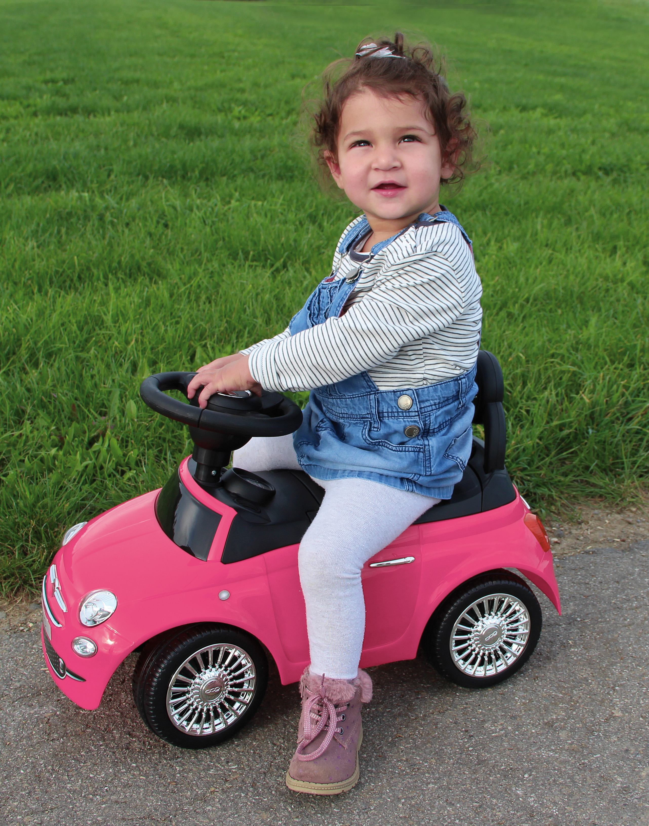 Ragazza su un'auto giocattolo rosa, che guida sull'asfalto, erba verde sullo sfondo, giornata di sole.