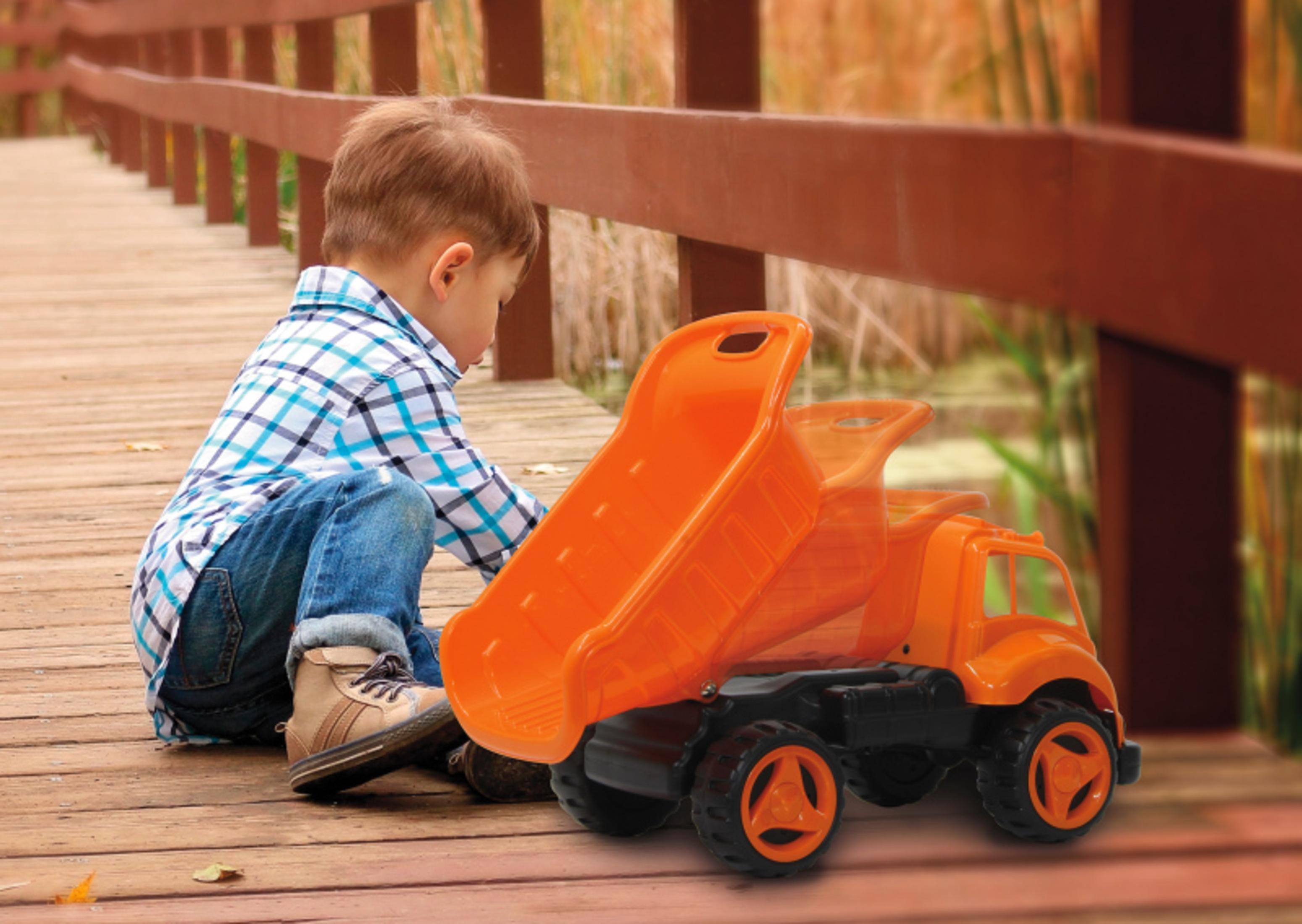 Ein Junge spielt mit einem orangen Muldenkipper-Spielzeug auf einer Holzbrücke. Hintergrund unscharf.