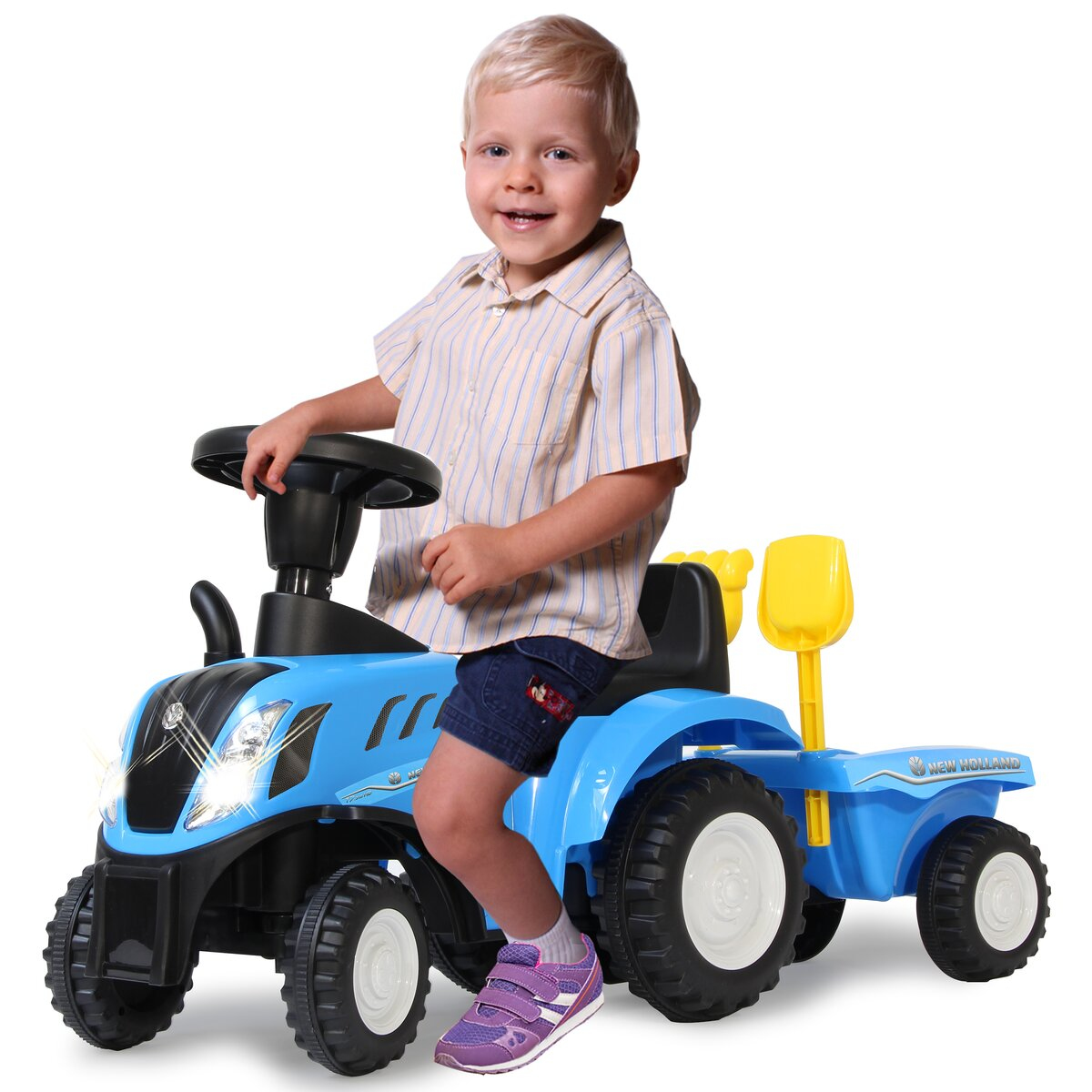 Niño en tractor de juguete azul con remolque. Marca New Holland, herramientas amarillas.