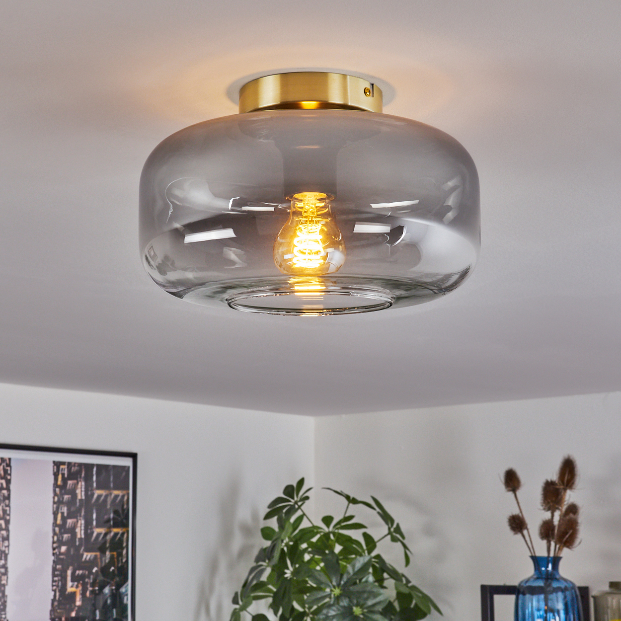 Eine Deckenlampe mit Glasschirm und goldenem Sockel. Eine leuchtende Glühbirne ist sichtbar. Pflanzen im Hintergrund.