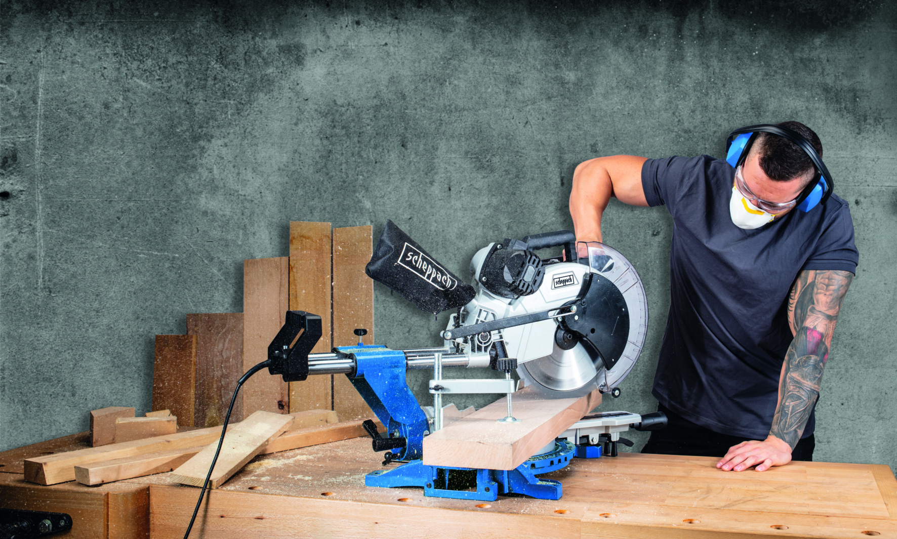 Man met veiligheidsuitrusting die een verstekzaag gebruikt om hout te zagen in een werkplaats, betonnen muur op de achtergrond.