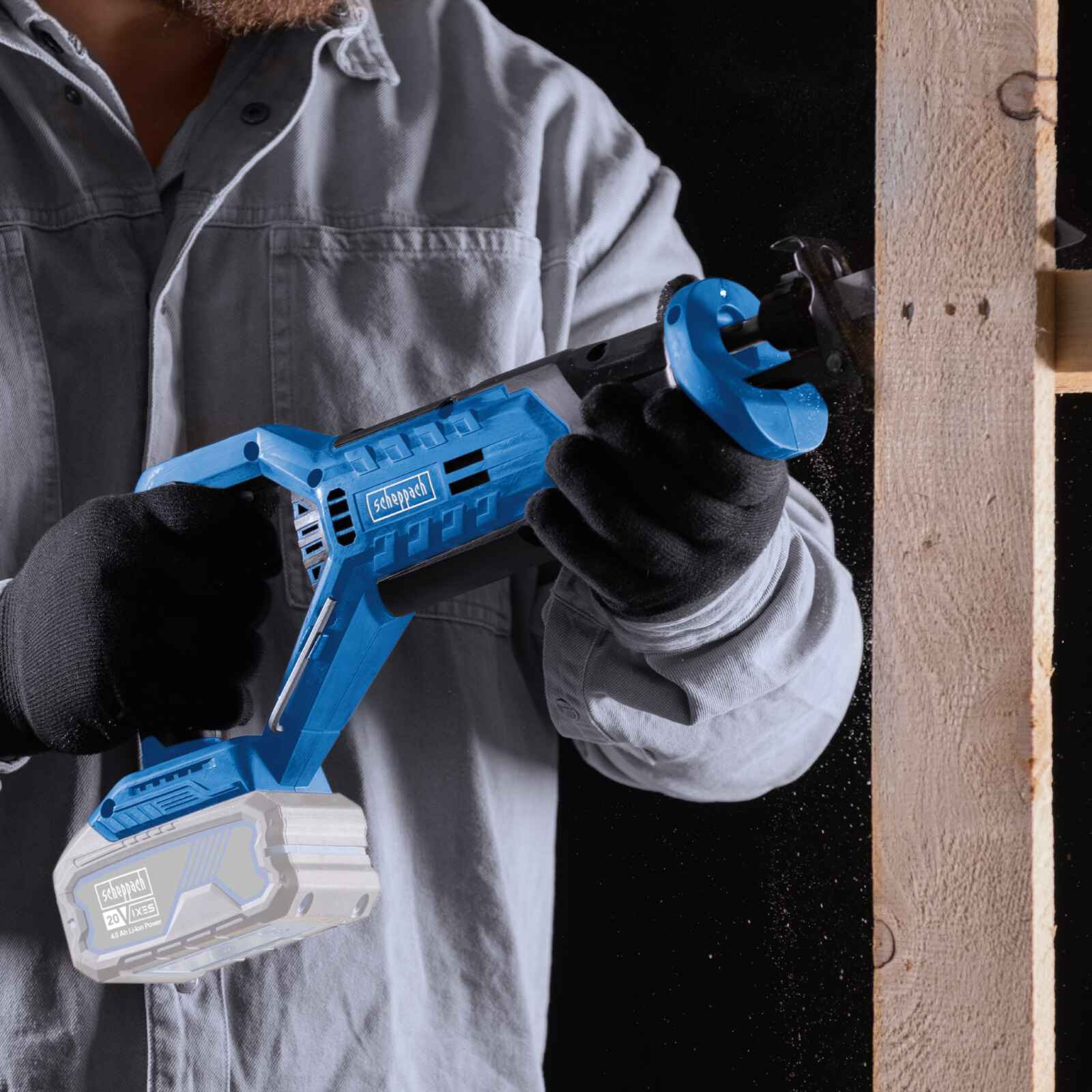 Een persoon snijdt een houten pallet met een blauwe reciprozaag. Ze dragen handschoenen in een donkere context.