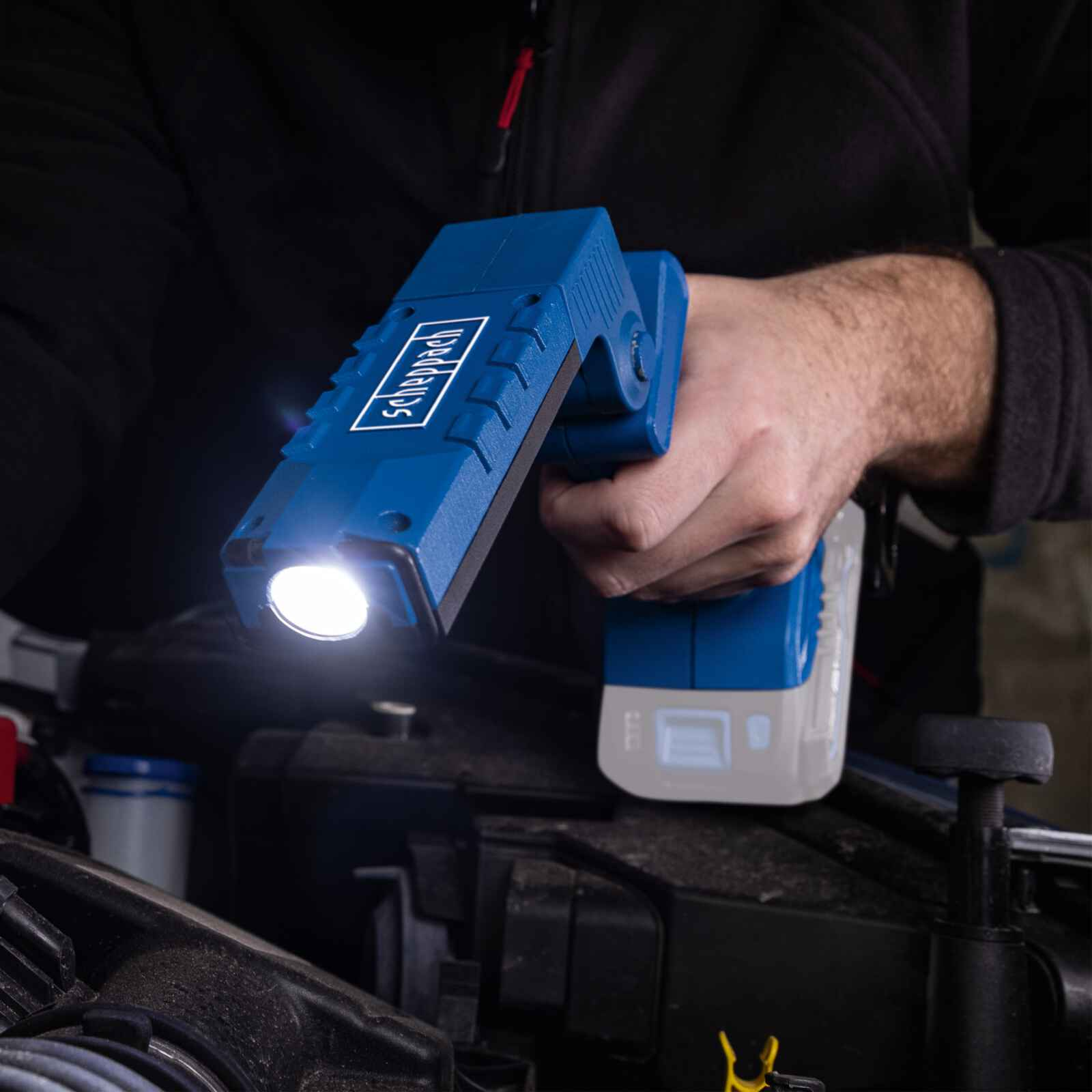 A person holding a blue Scheppach flashlight over a car engine bay.