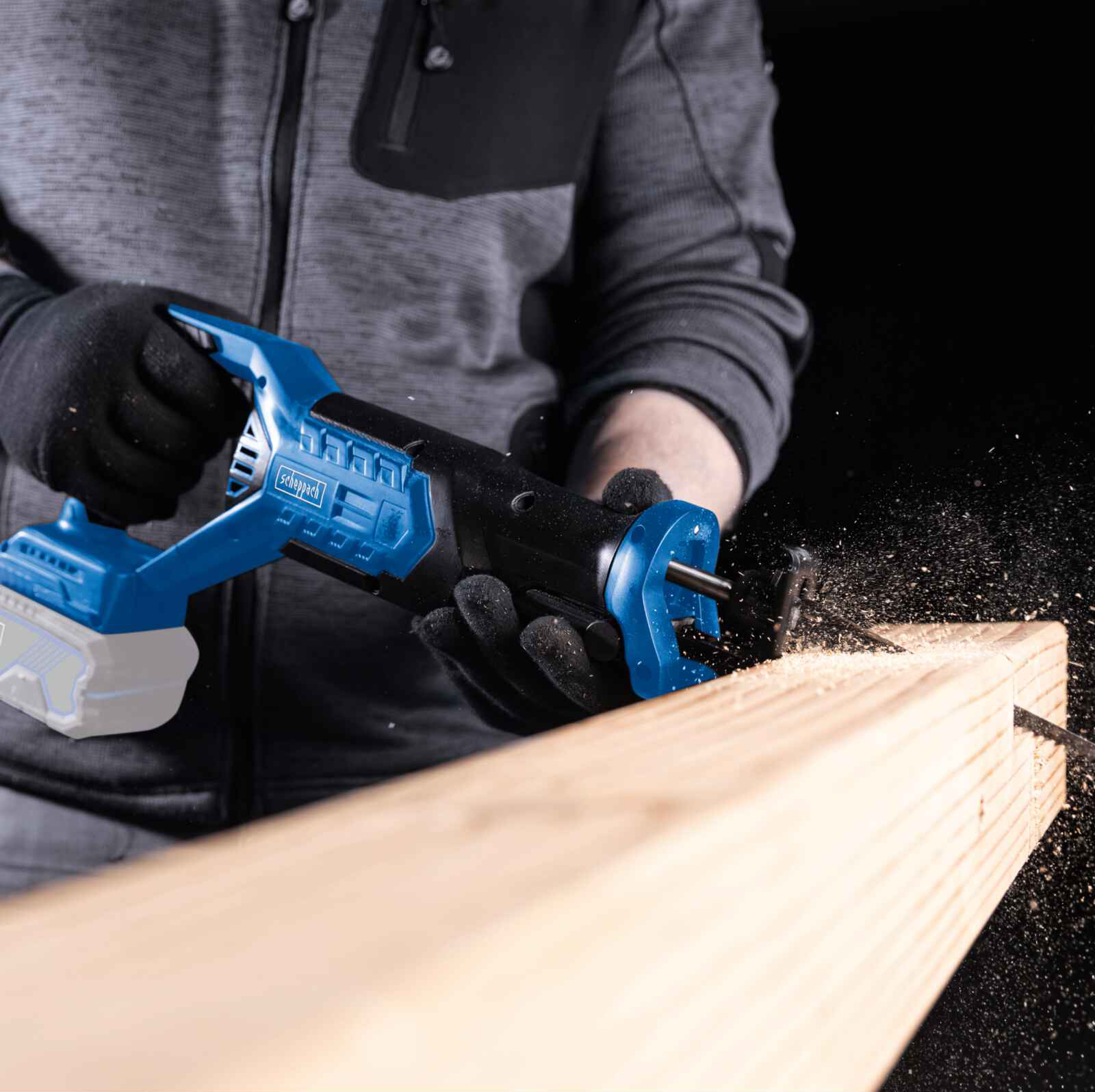 Person in grauem Hemd und schwarzen Handschuhen benutzt eine blaue Säbelsäge auf einem Holzbrett.