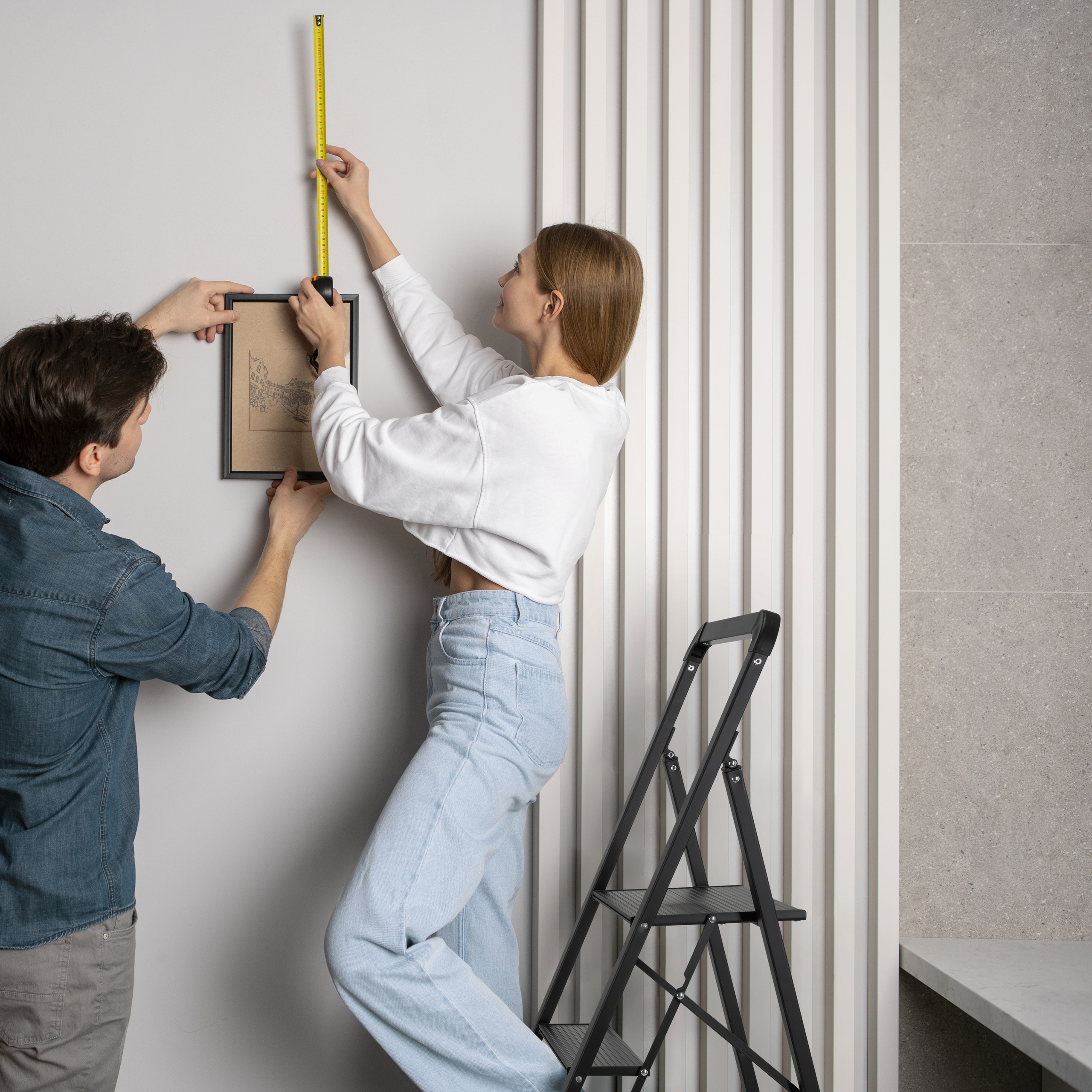 Ein Mann und eine Frau hängen ein Bild an die Wand. Die Frau misst mit einem Maßband und steht auf einer Leiter.