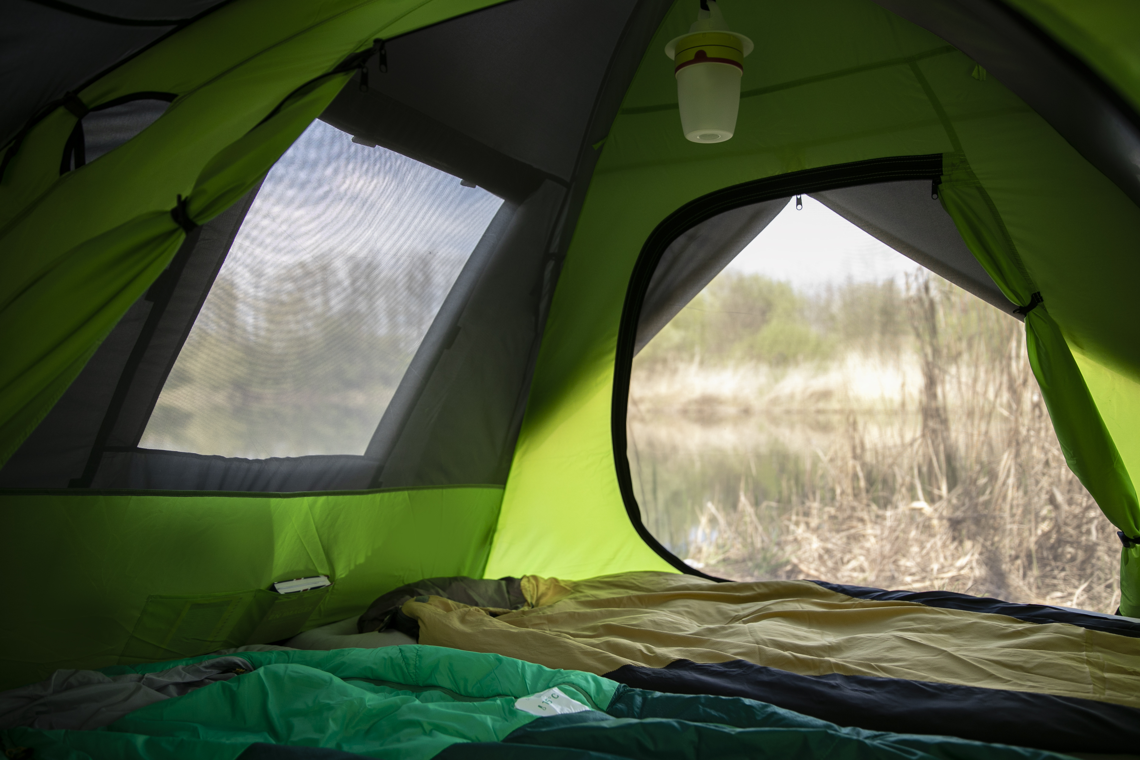 Blick von innen aus einem grünen Zelt auf einen See durch ein Netzfenster. Schlafsack innen.