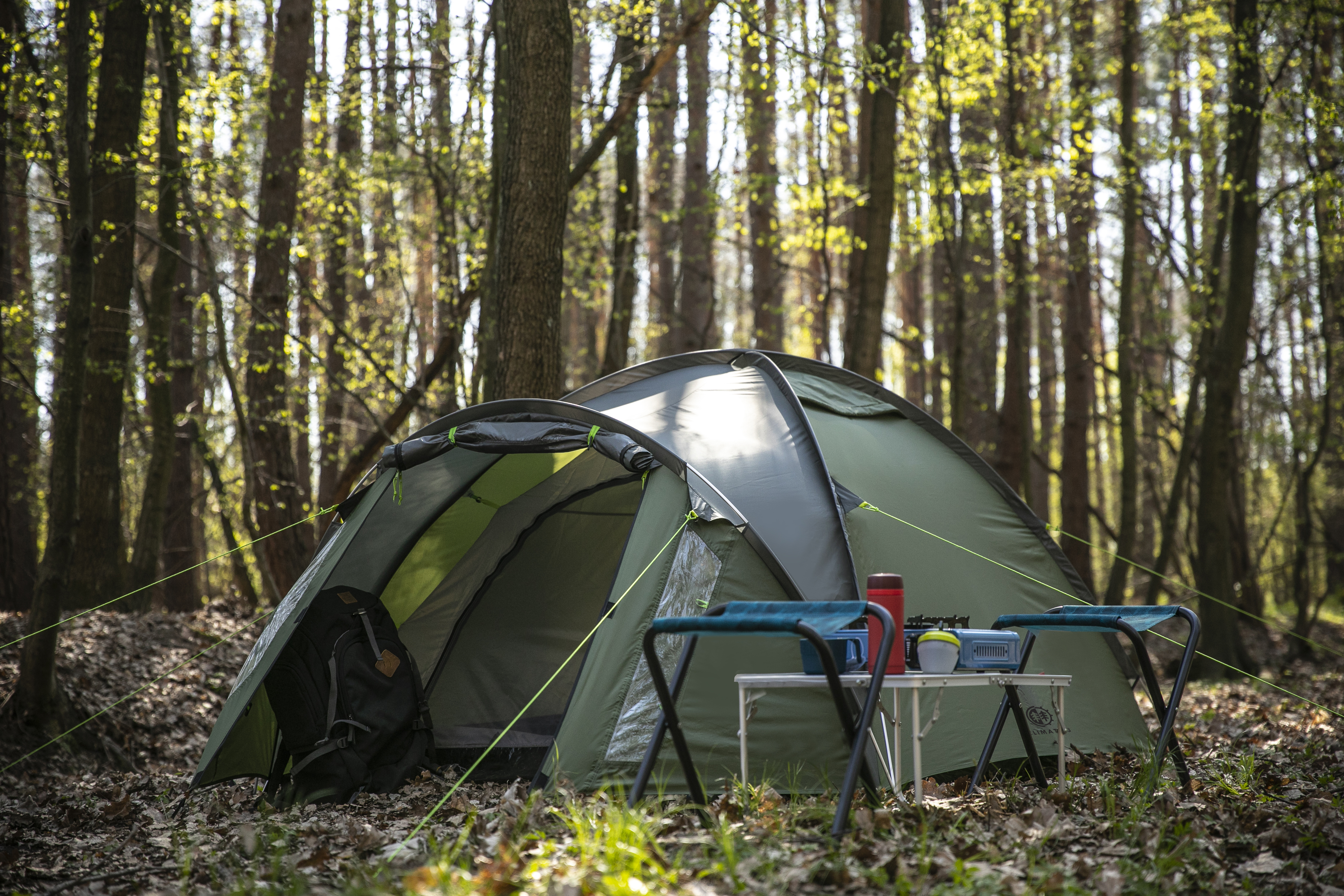 Grünes Zelt im Wald aufgestellt. Tisch, Campingstühle und Rucksack sichtbar. Bäume und Sonnenlicht.