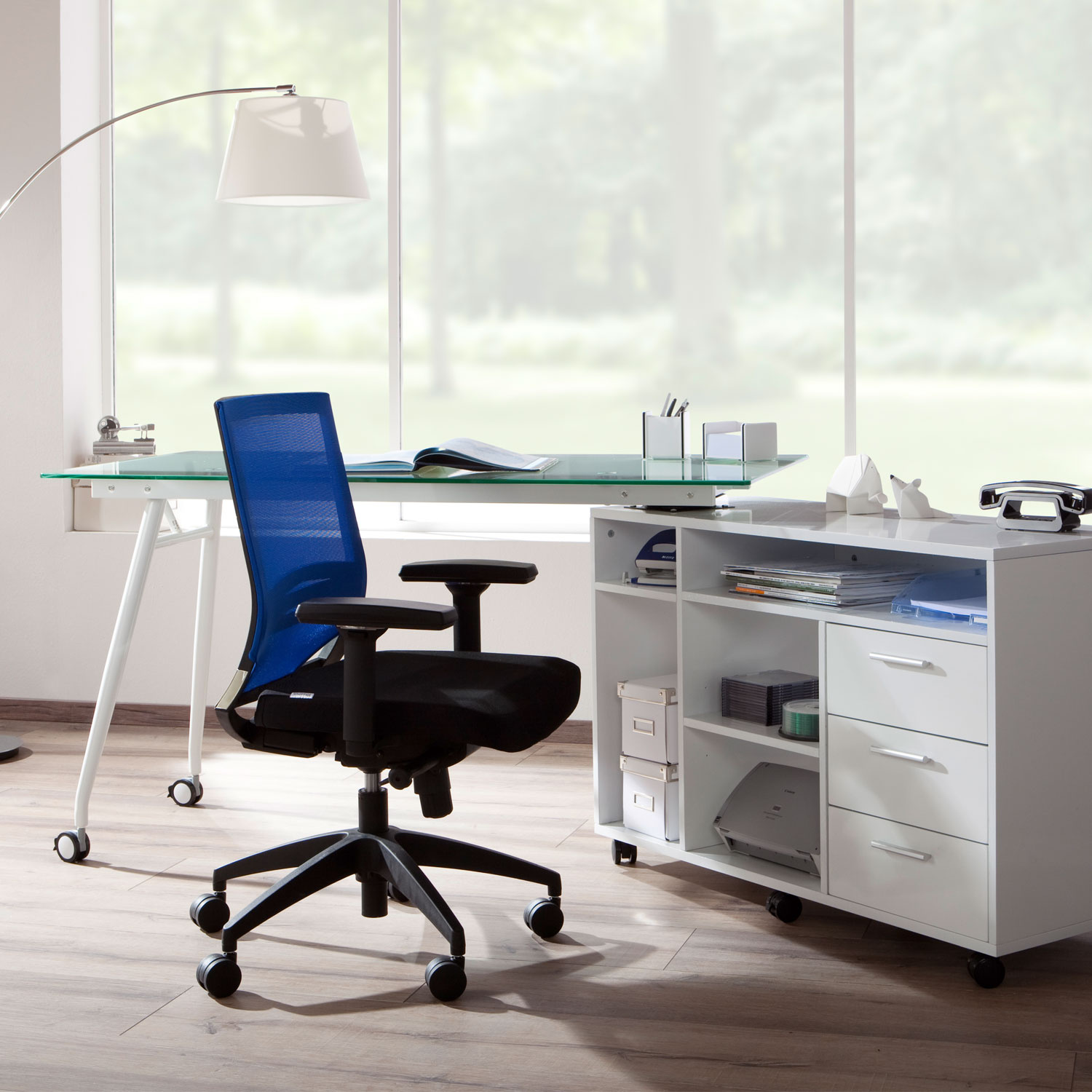 Bürotisch-Setup mit blauem Netzrückenstuhl, weißem Schreibtisch und Schrank, neben einem Fenster und einer Stehlampe.