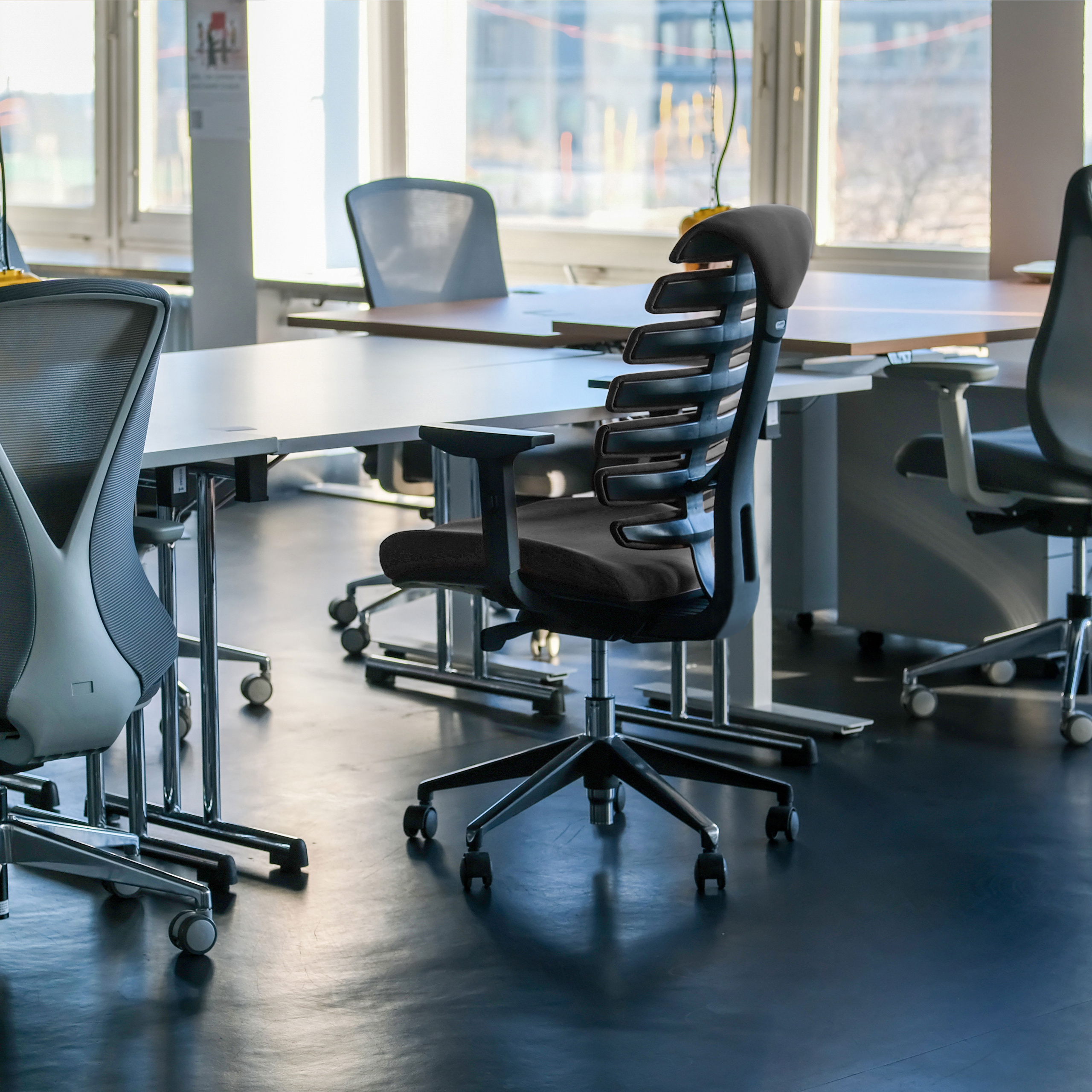 Büro mit modernen schwarzen Stühlen, langen weißen Tischen und großen Fenstern mit Sonnenlicht.