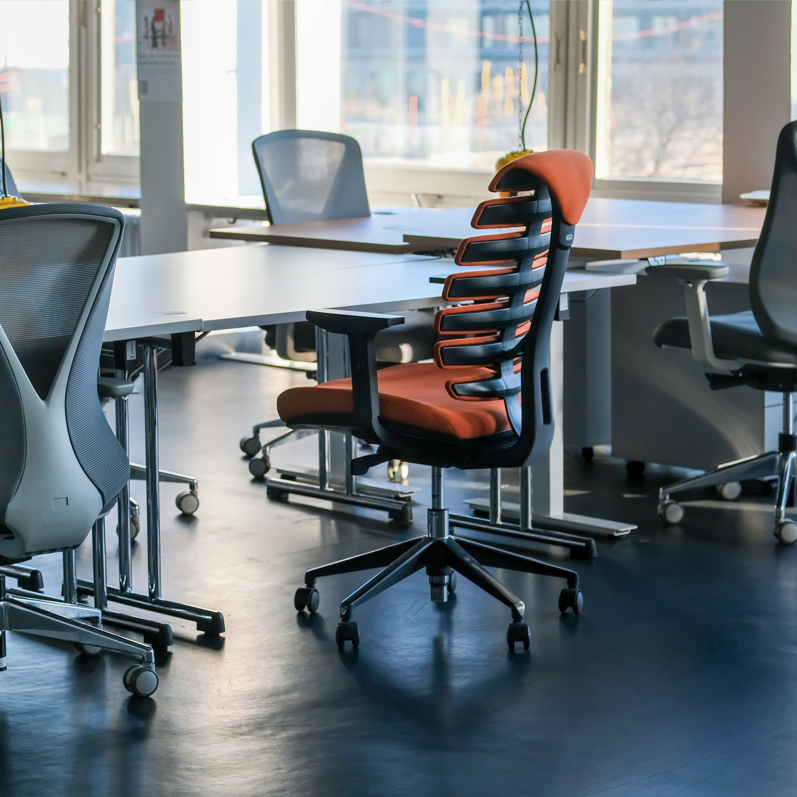 Büro mit Stühlen, Schreibtischen und großen Fenstern. Ein orangefarbener und schwarzer Stuhl steht im Mittelpunkt.