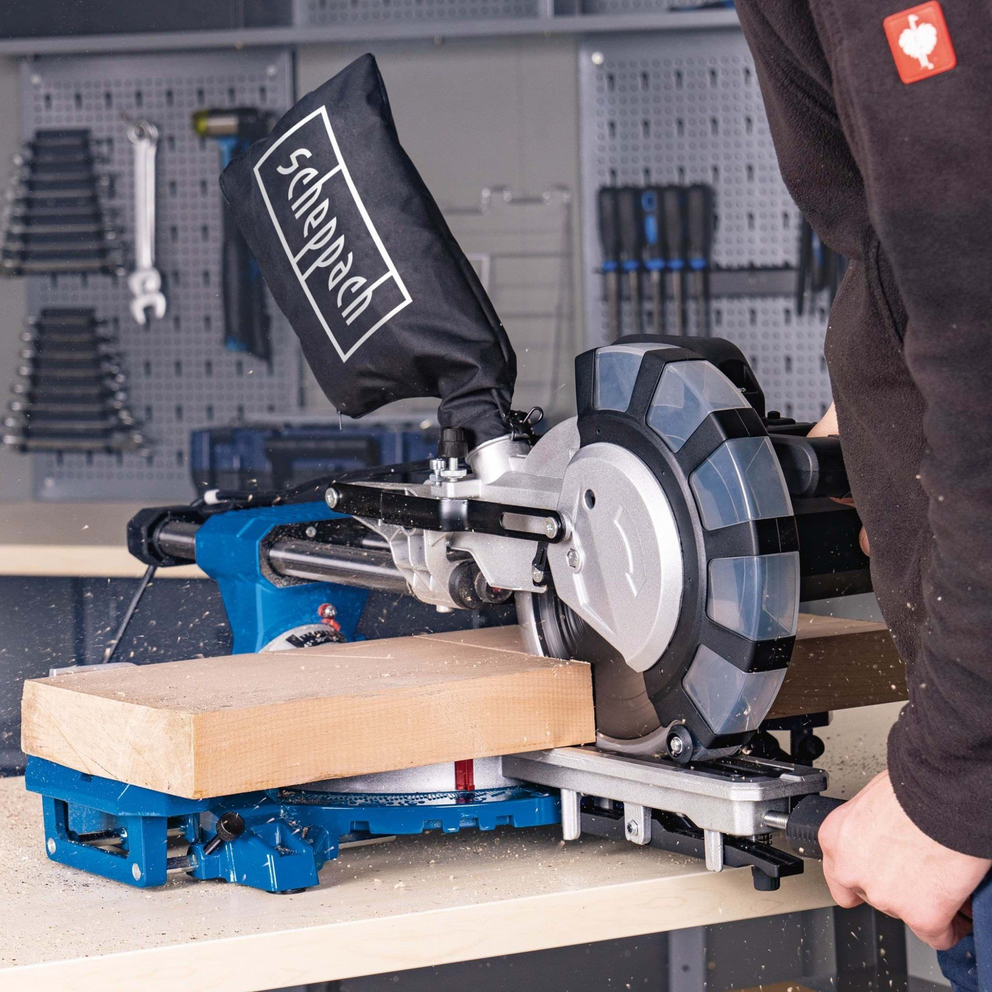 Man die hout zaagt met een blauw-zwarte zaagmachine op een werkbank, grijze muur.