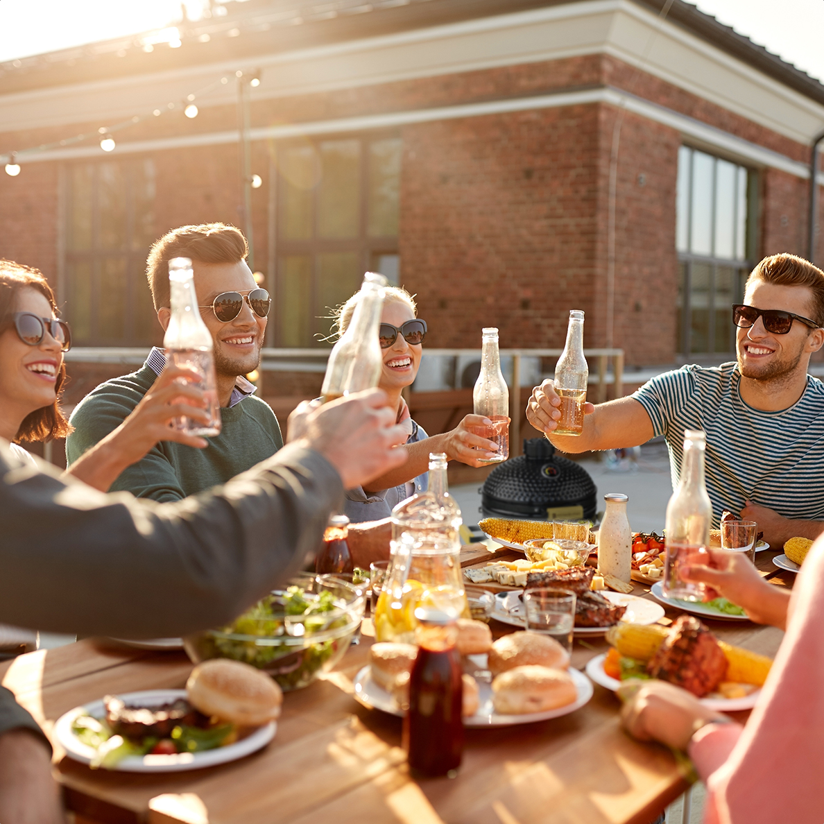 Des personnes trinquent avec des bouteilles de bière lors d'un barbecue, avec de la nourriture et un grill visibles.