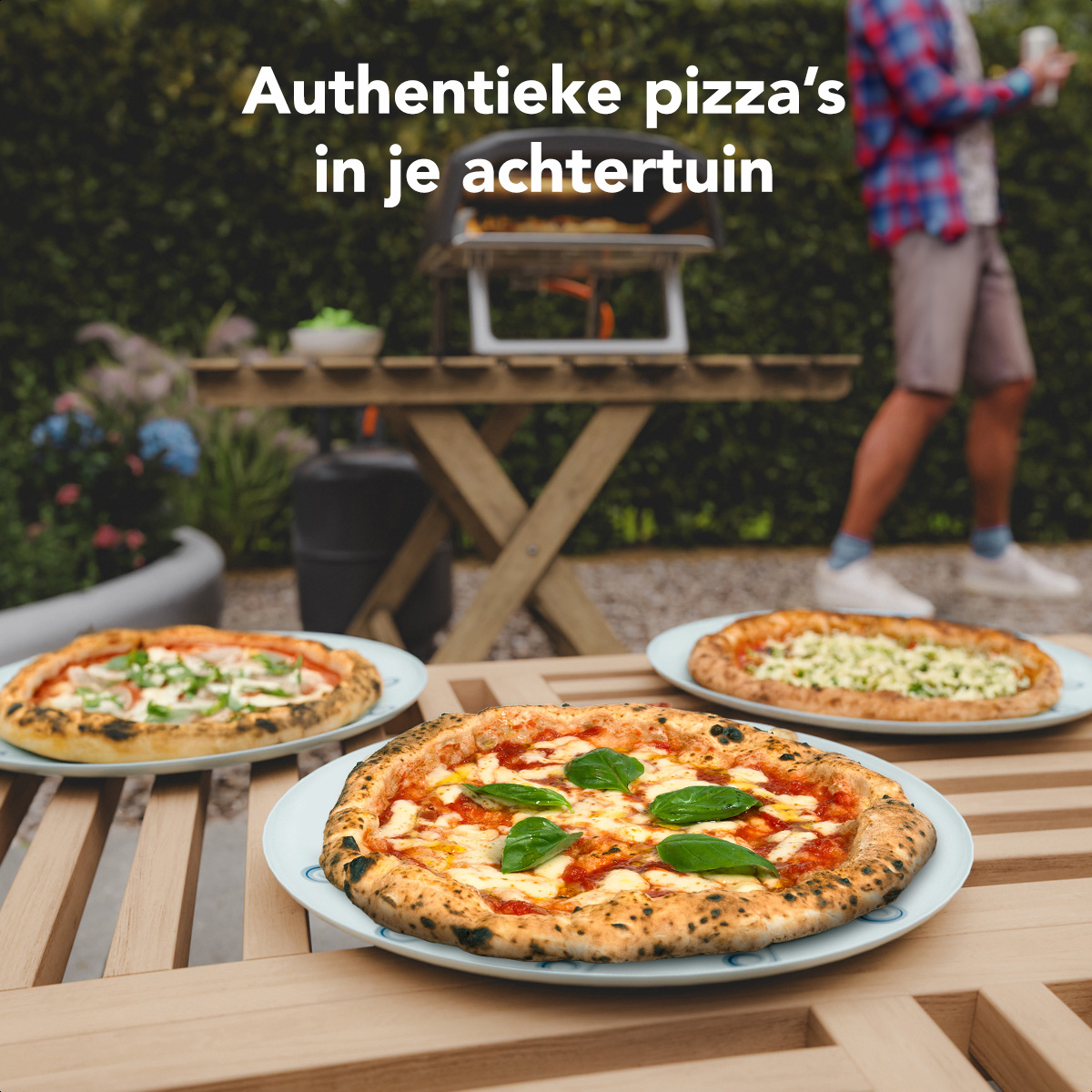 Three pizzas on plates on a wooden table. Pizza oven in the background in a garden setting.