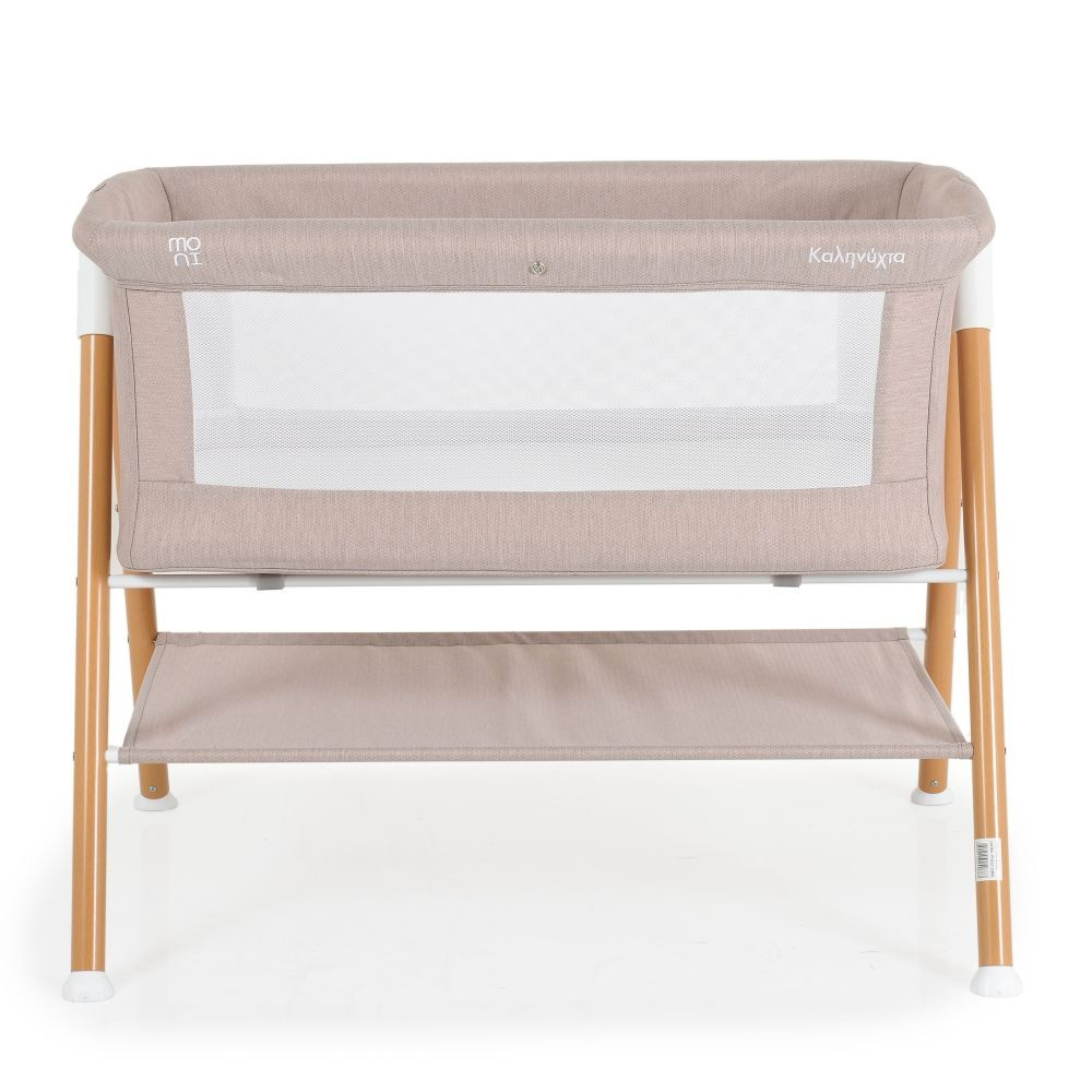 Una cuna beige para bebés con un lado de malla blanco. Patas de madera y un estante de almacenamiento. Fondo blanco.