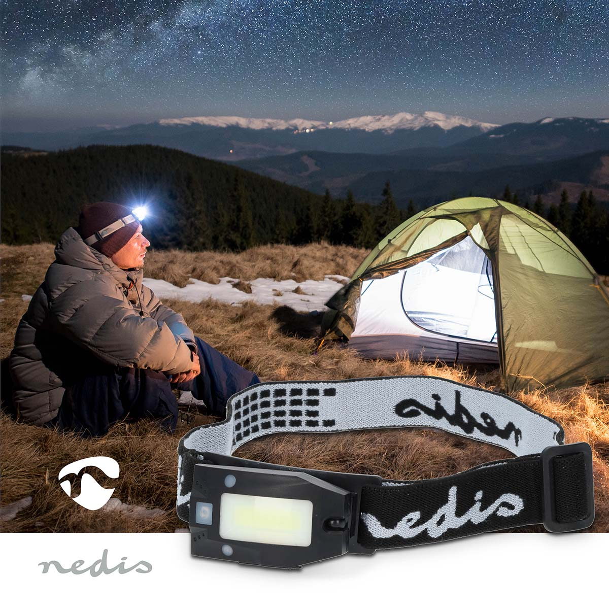 Man in a campsite wearing a headlamp, tent and mountains in the background.