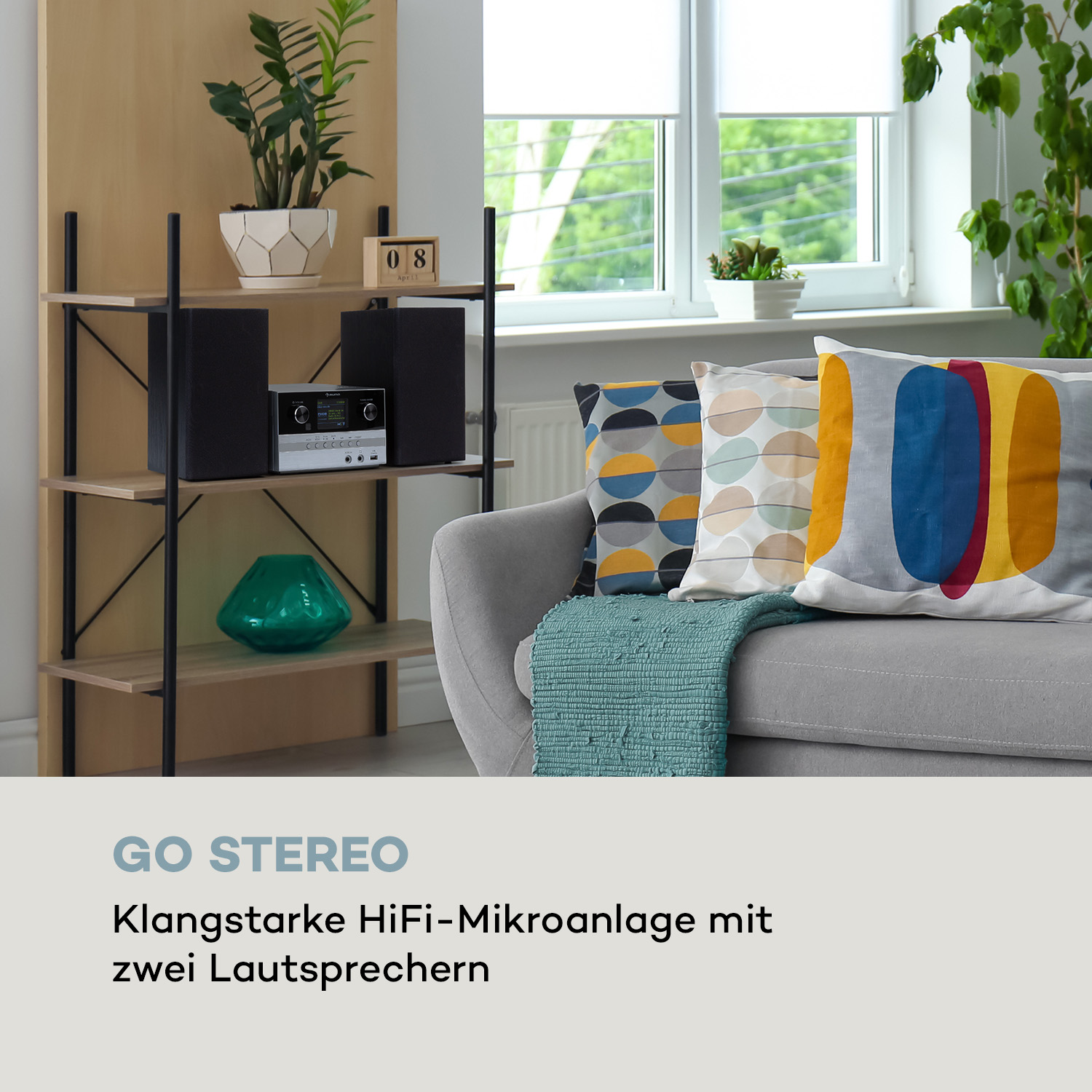 Wohnzimmer mit Stereoanlage, Büchern, Fenster und Pflanzen. Graues Sofa mit bunten Kissen.