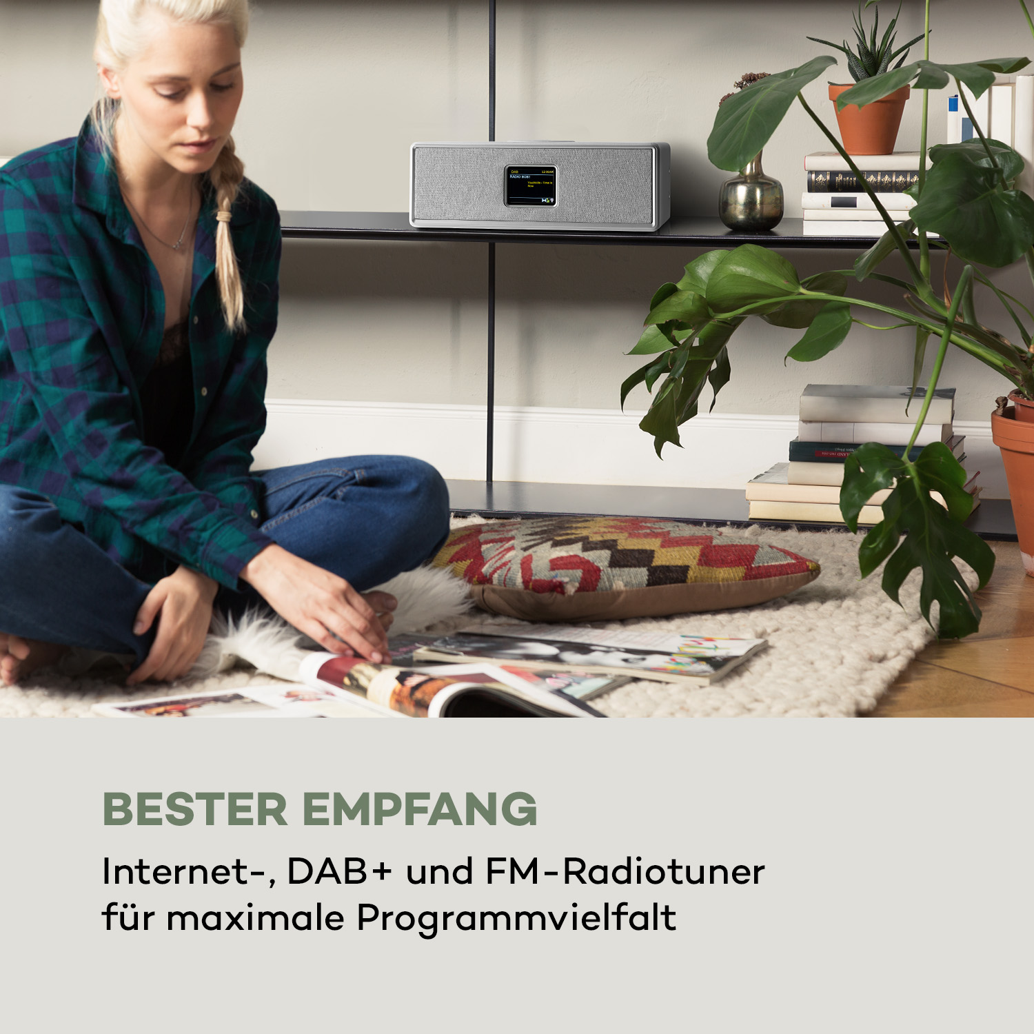 Frau sitzt neben einem Regal mit Radio und Pflanzen und liest eine Zeitschrift.