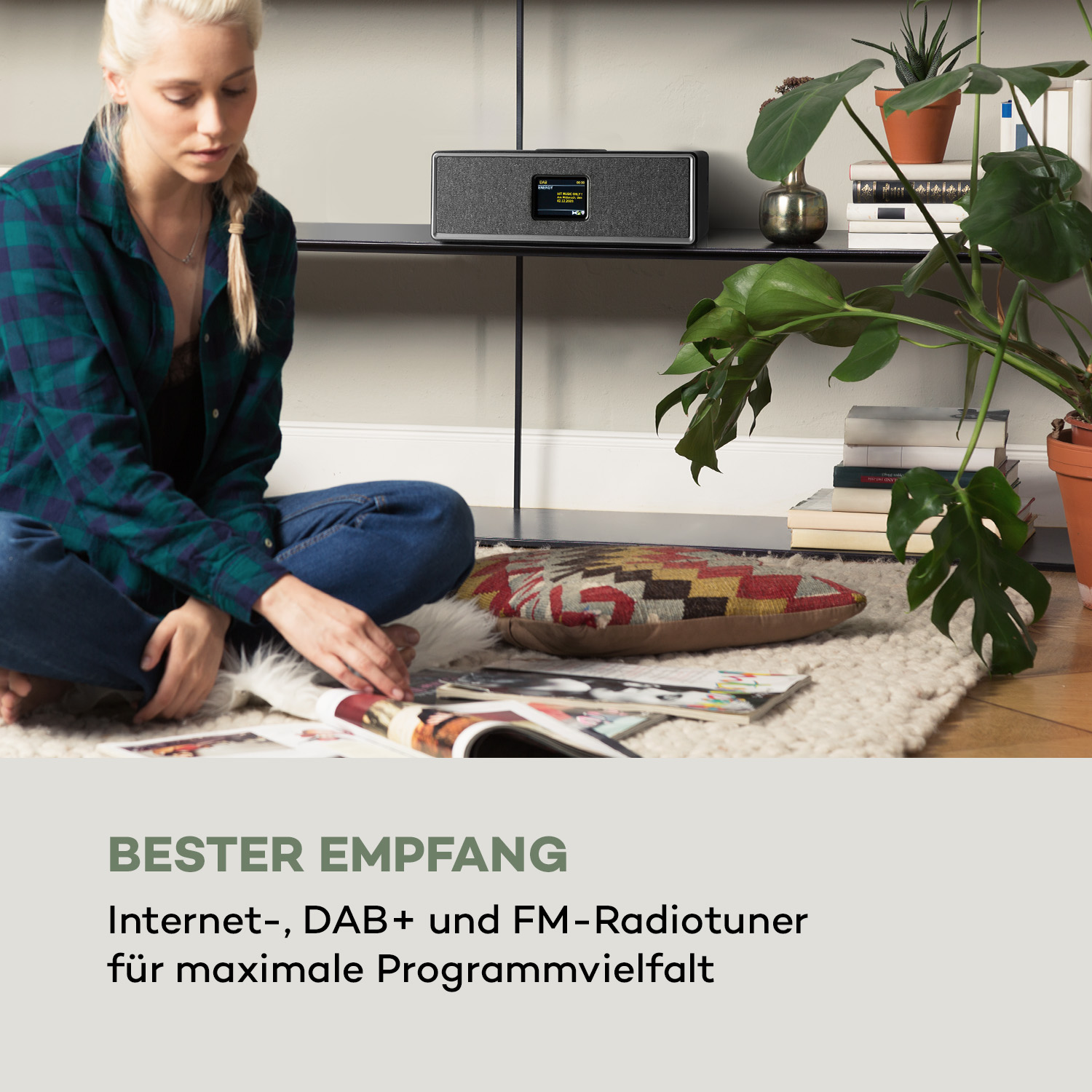 Eine Frau sitzt in der Nähe eines Radios und liest. Der Raum umfasst Pflanzen und einen Teppich. Radio hat einen Bildschirm.