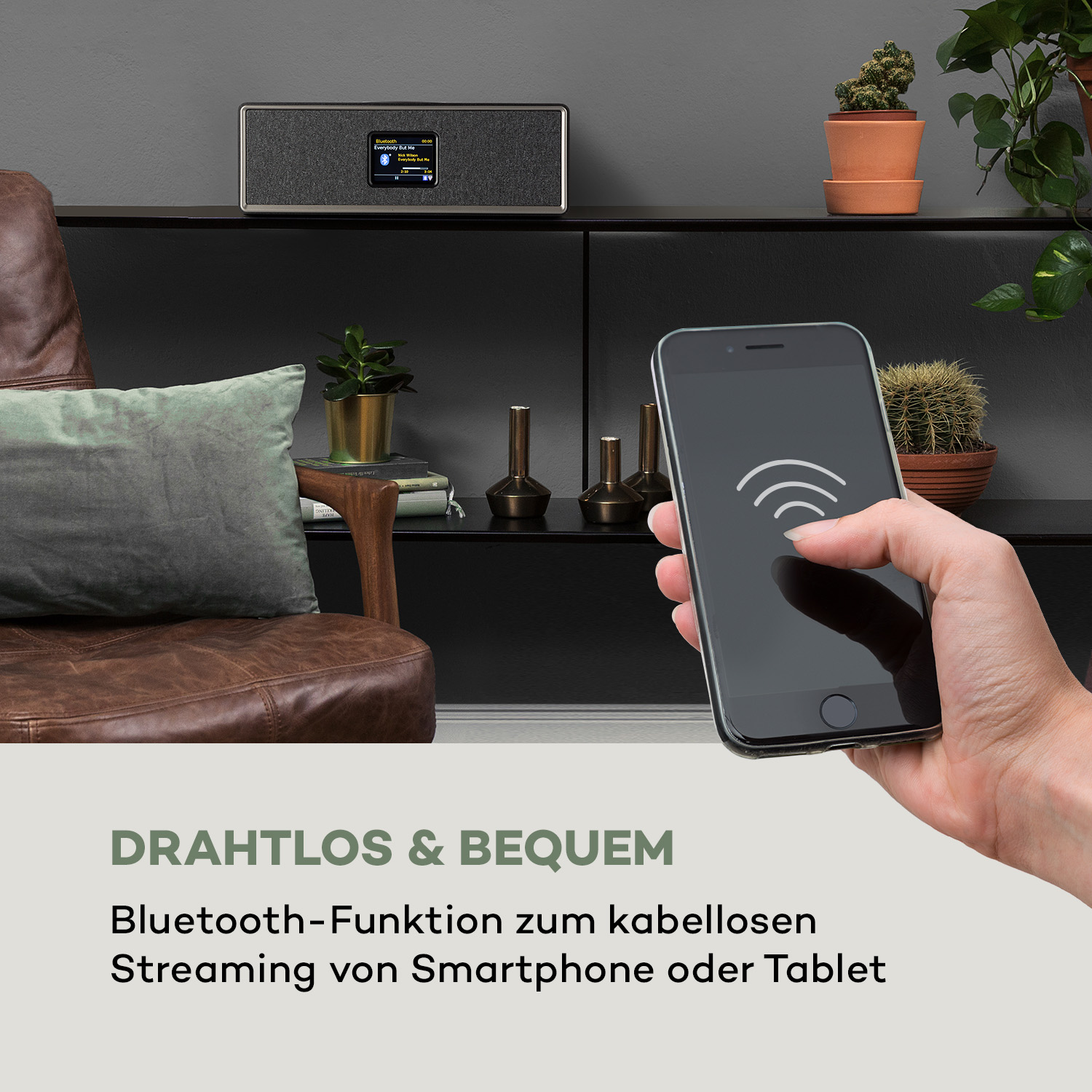 Eine Hand hält ein Smartphone, das sich mit einem schwarzen Radio verbindet. Pflanzen und ein Ledersessel im Hintergrund.