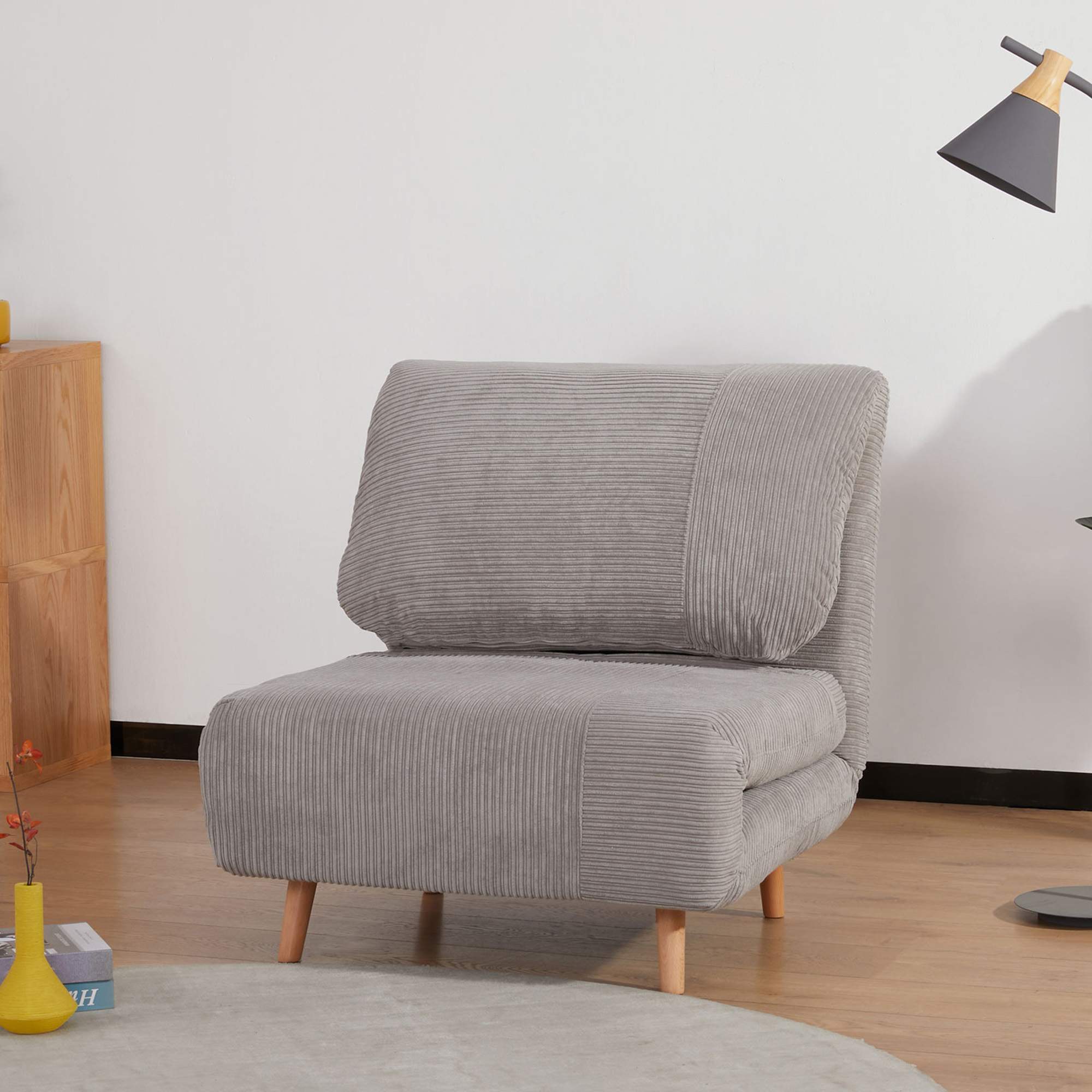 Una sedia in velluto a coste grigio è in una stanza. Ha gambe in legno ed è vicino a una lampada e a un tappeto.
