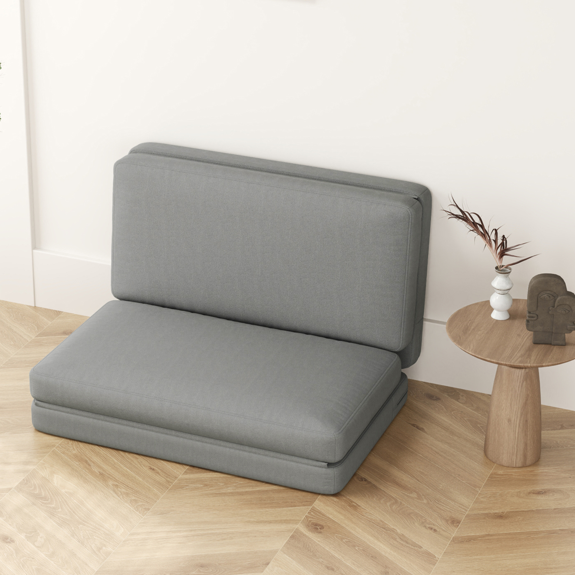 Graues Klappsofa, zu einem Sitz zusammengeklappt. Ein kleiner Tisch und Dekorationen daneben.
