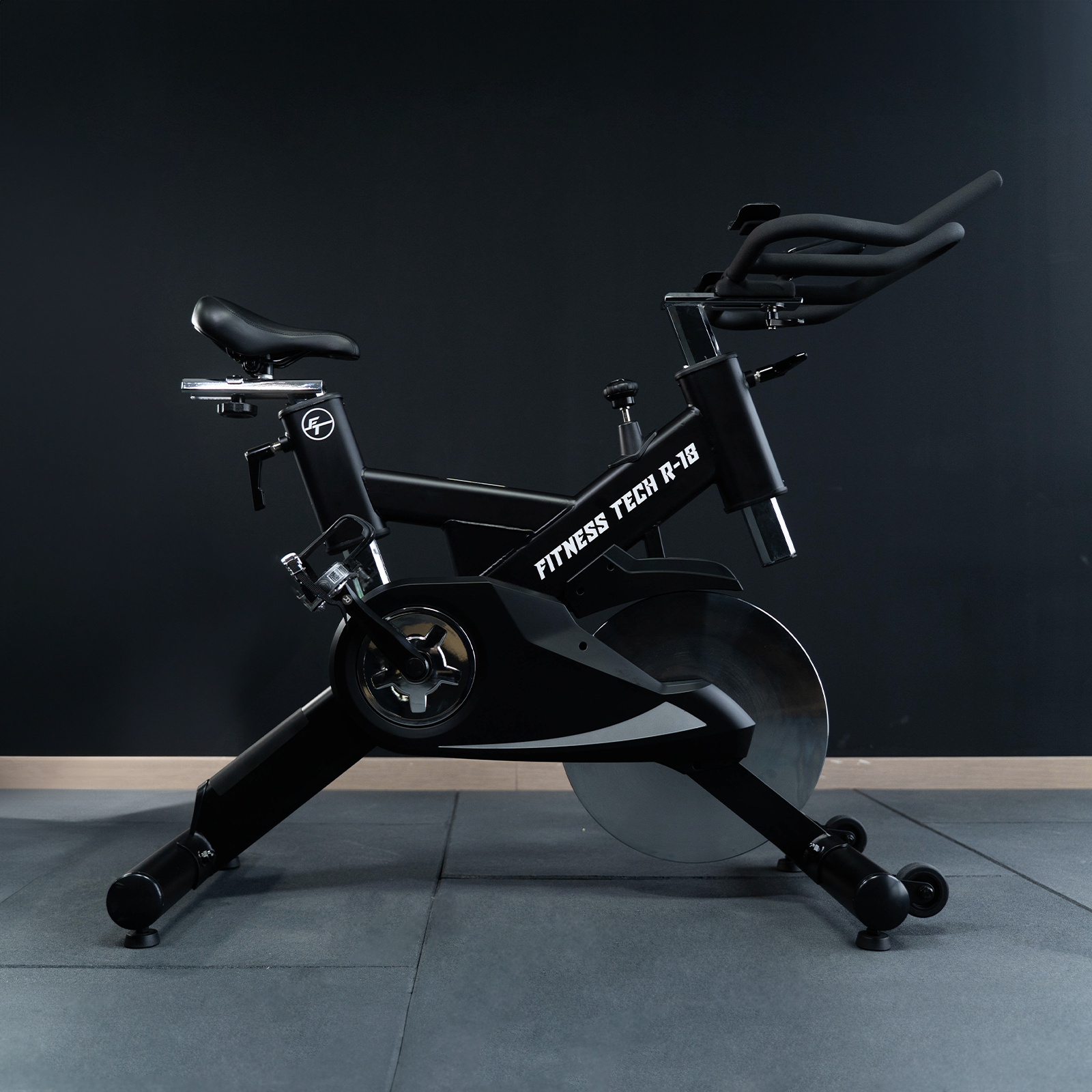 Bicicleta de ejercicio negra, vista lateral, Fitness Tech R-18, suelo gris, fondo oscuro.