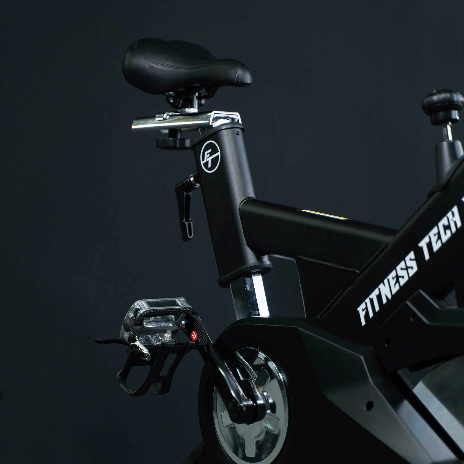 Detalle de bicicleta de ejercicio negra, asiento, pedal, logo Fitness Tech, fondo negro.