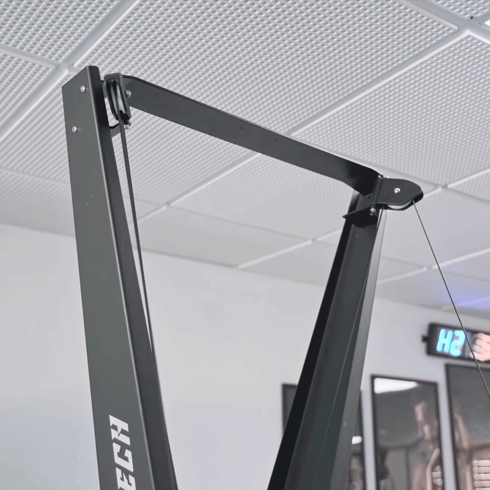 Máquina de gimnasio negra y gris, con sistema de cable, contra pared blanca con reloj.