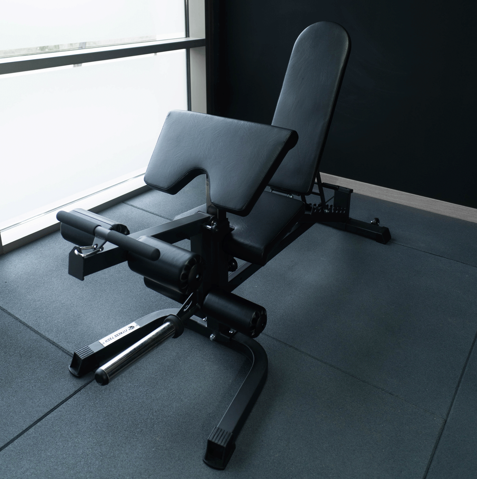 Banco de gimnasio negro con reposabrazos. Cerca de una ventana, con pared negra y suelo gris.