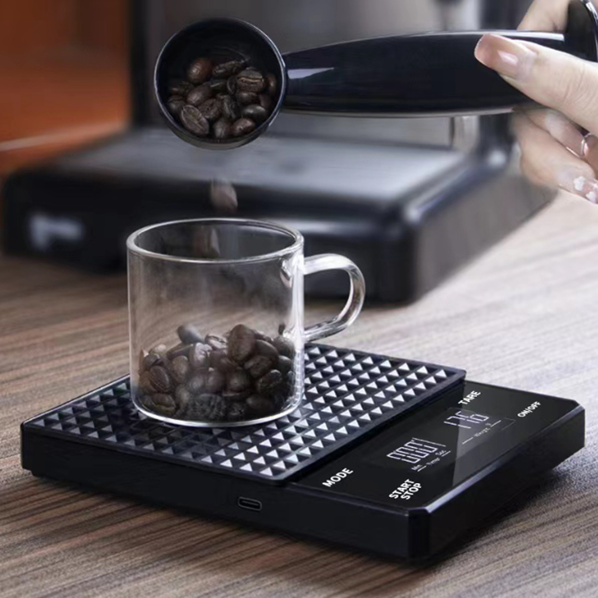 Schwarze Küchenwaage mit Kaffeebohnen in einem Glas. Eine Schaufel Bohnen wird in das Glas gegossen. Das Display zeigt das Gewicht.